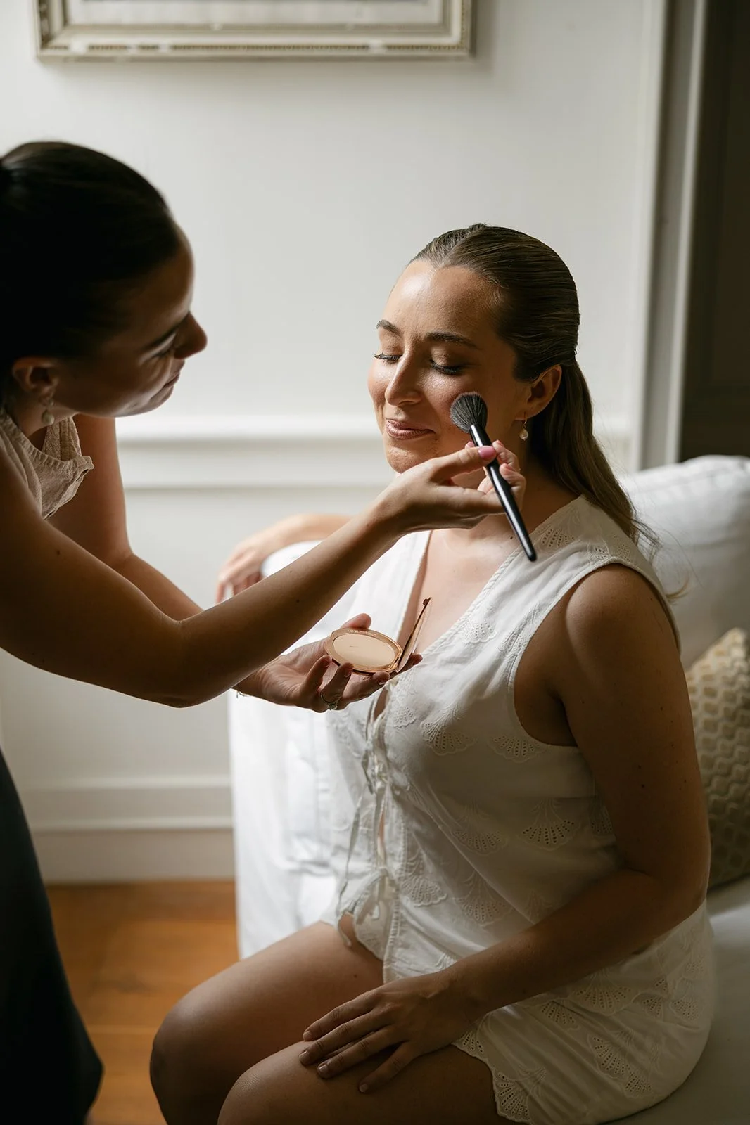 A makeup artist applies blush to seated woman in a white lace outfit in a softly lit room, creating a calm, elegant beauty or bridal preparation scene.