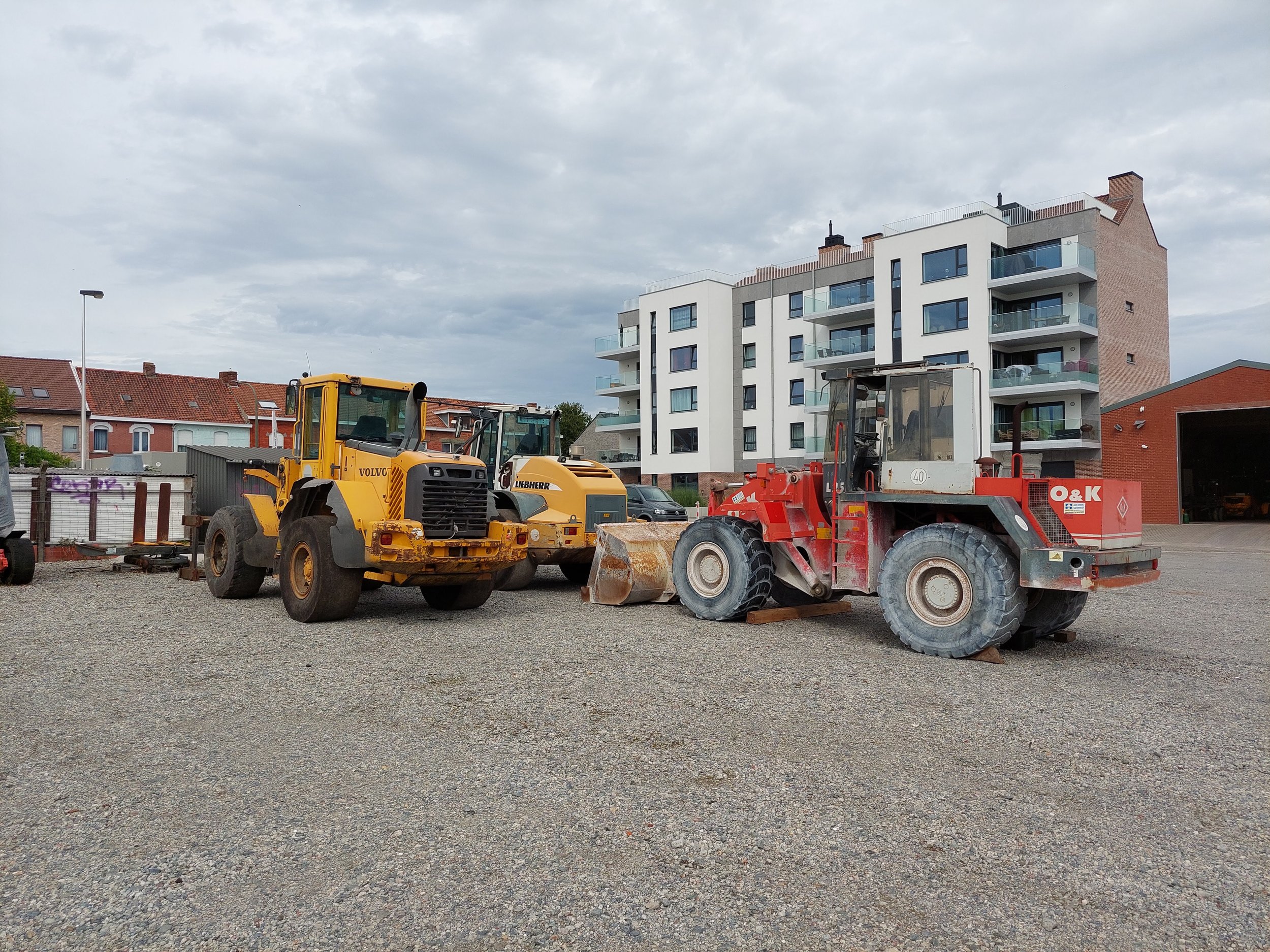 Twee zware bouwvoertuigen, een gele Volvo en een rode O&K, staan op een onbestemde bouwplaats met een gravel oppervlak, met in de achtergrond een modern wit appartementencomplex en een paar kleinere gebouwen onder een bewolkte hemel.