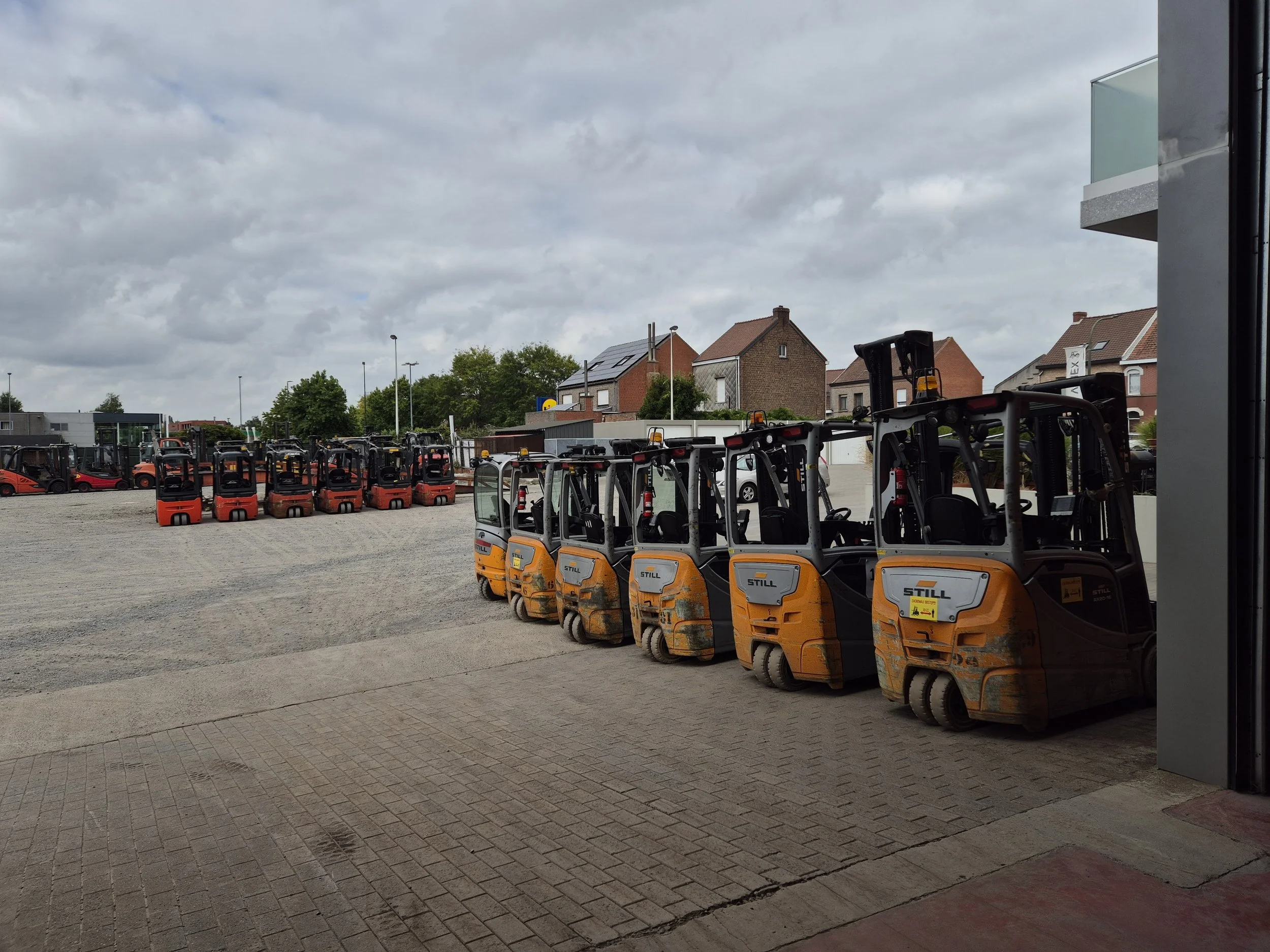 Rijen elektrische heftrucks en logistieke voertuigen buiten op een parkeerplaats met huizen op de achtergrond onder een bewolkte lucht.