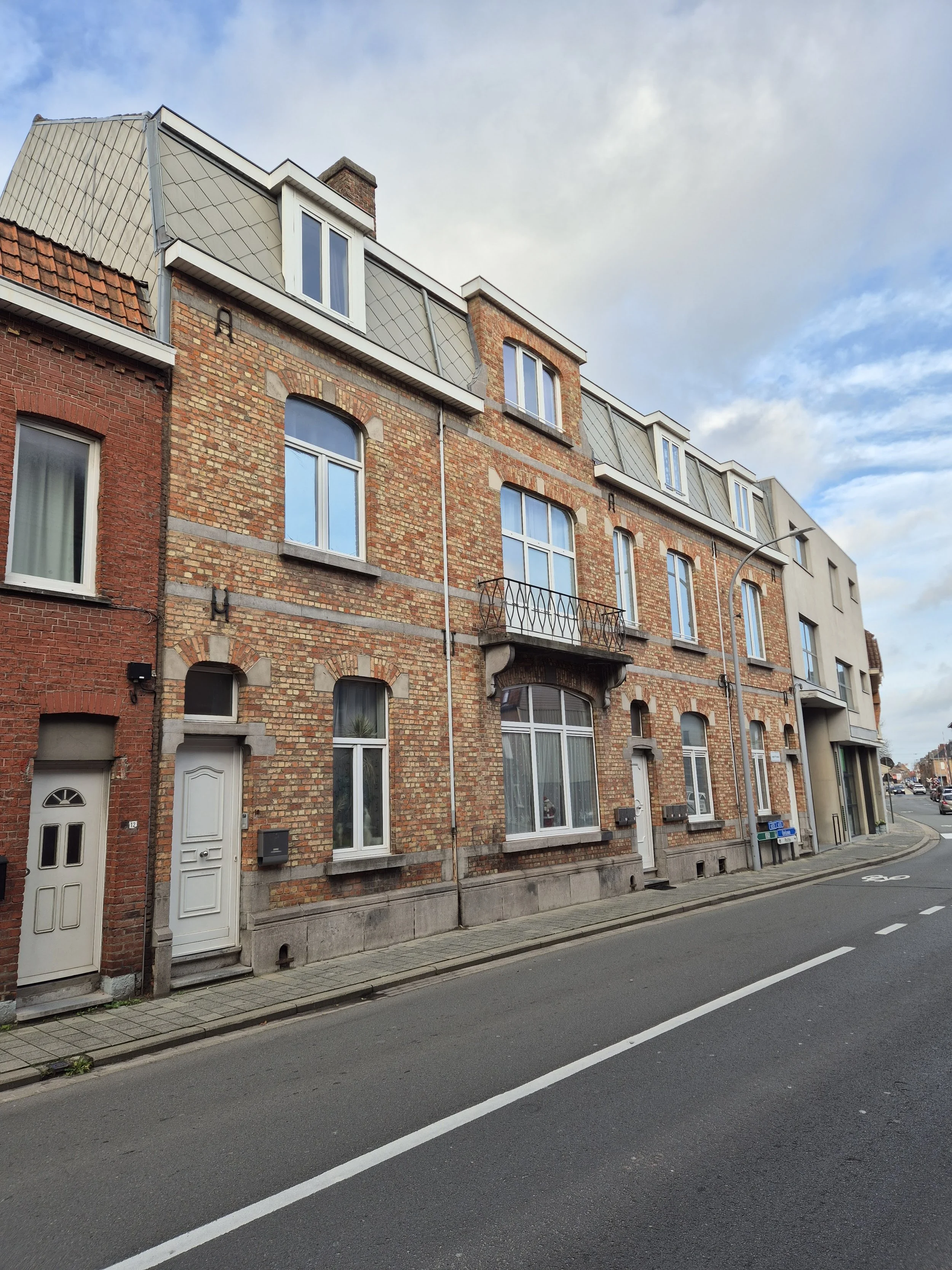 Rijhuis met rode bakstenen gevel, verschillende ramen, een kleine balkon en een moderne bovenverdieping, langs een straat met fietspad en wegmarkeringen.