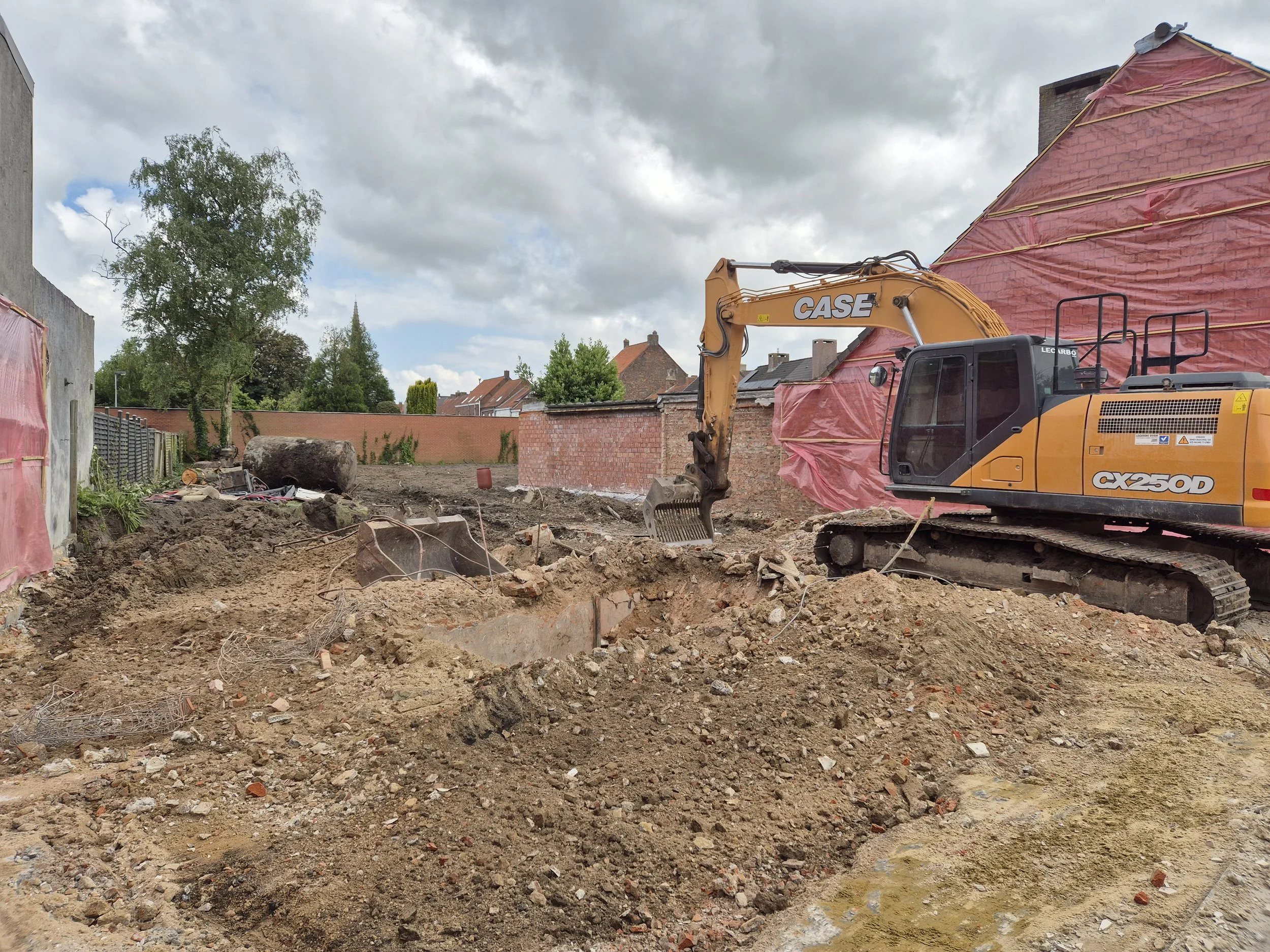 Bouwplaats met een gele case CX250D graafmachine op een ontginningsplek met aarde en stenen, omgeven door een muur en gebouwen met rood dak en een grote boom op de achtergrond.