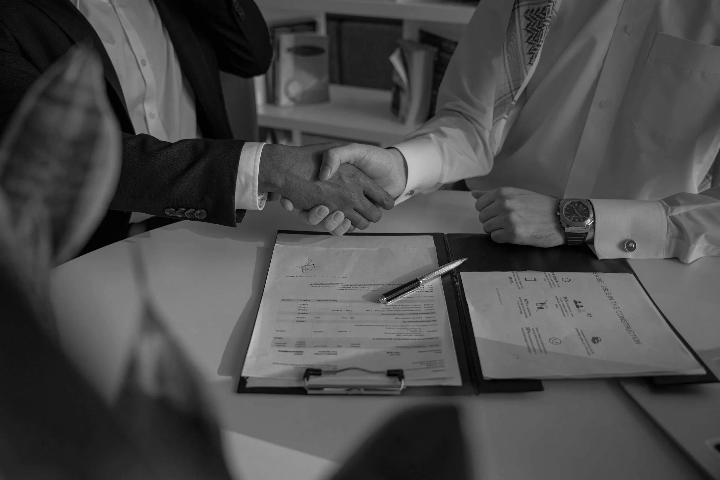 Twee mensen schudden elkaar de hand tijdens een zakelijke bijeenkomst, met documenten en een pen op tafel.