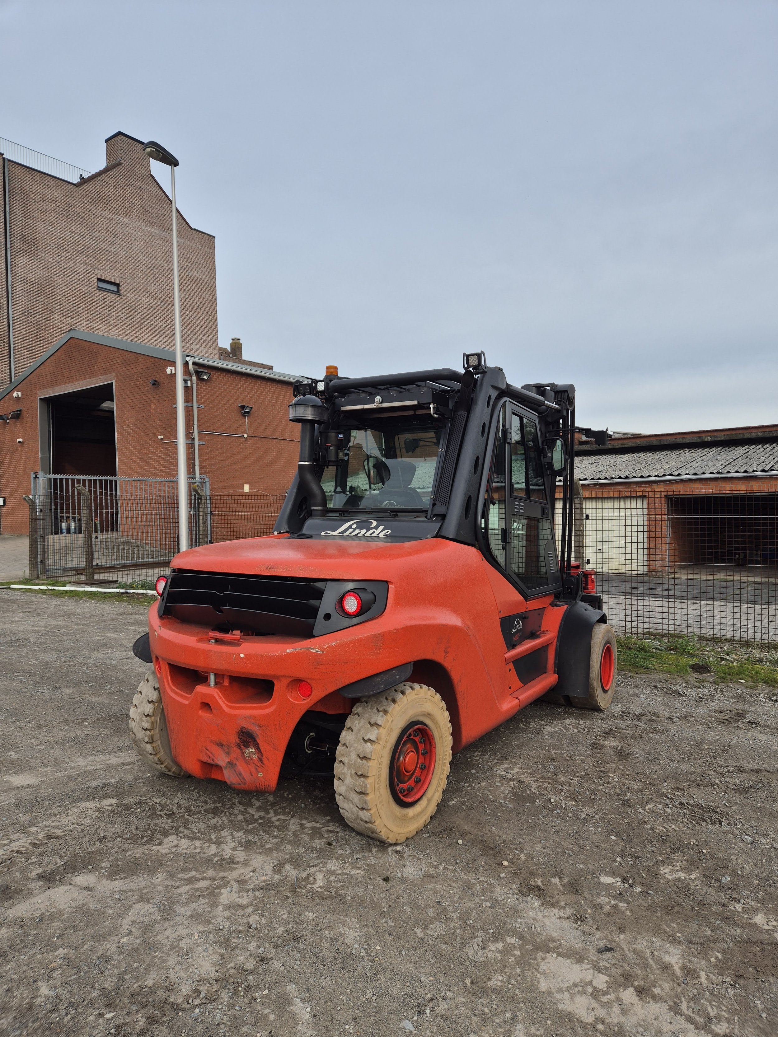 Een rode heftruck met vier stevige banden op een onverharde grond, met een gebouw en een hek op de achtergrond.