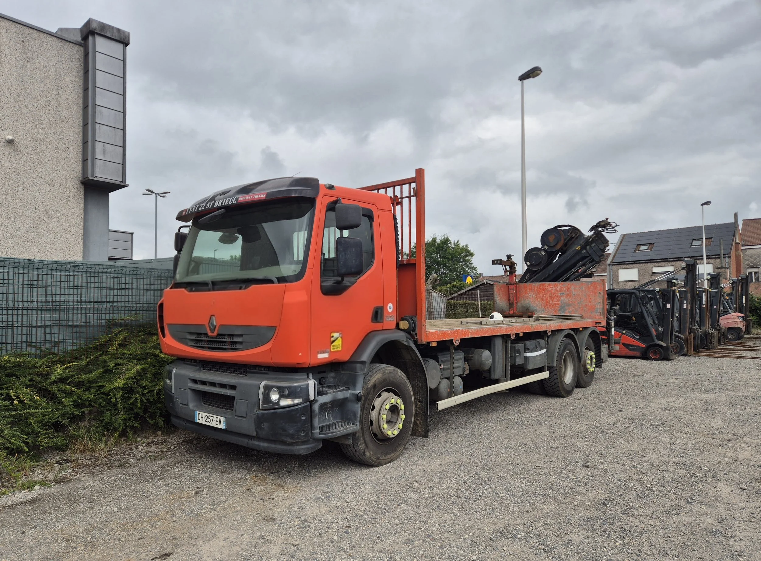 Een rode Renault vrachtwagen met open laadbak, geladen met stroomkabels en apparatuur, geparkeerd op een grindterrein met een rij vorkliften en gebouwen op de achtergrond onder een bewolkte hemel.