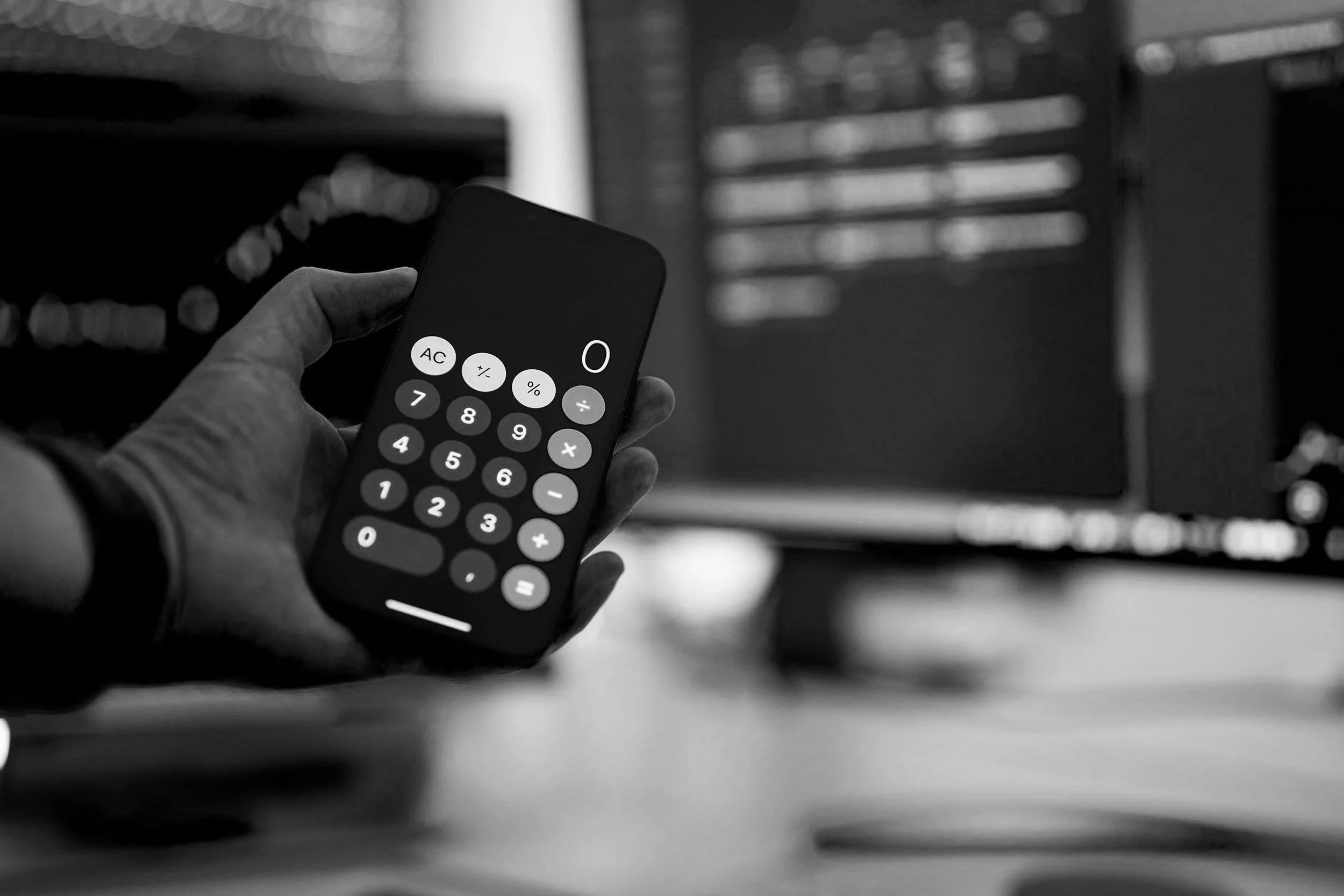 Een hand houdt een smartphone met een rekenmachine-app geopend, zwart-wit foto, op een bureau met een monitor in de achtergrond.