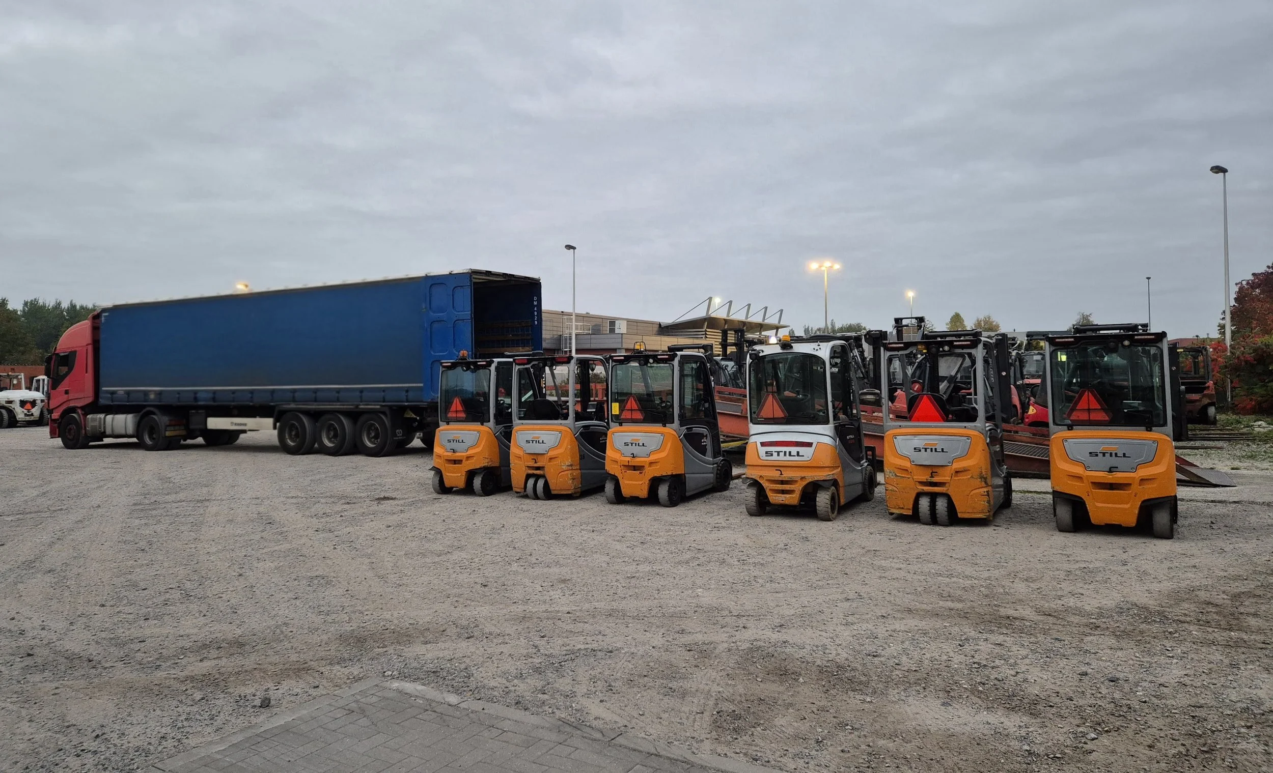 Verschillende kleine gele heftrucks en een grote blauwe vrachtwagen geparkeerd op een onherbergzaam terrein onder een bewolkte hemel.