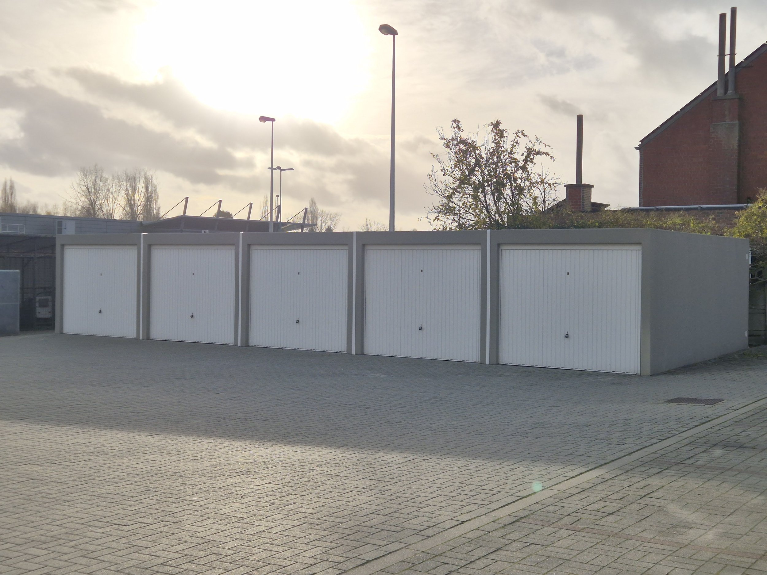 Een rij witte garagekooien met een betonnen muur erachter, onder een bewolkte hemel en met straatverlichting en bomen op de achtergrond.
