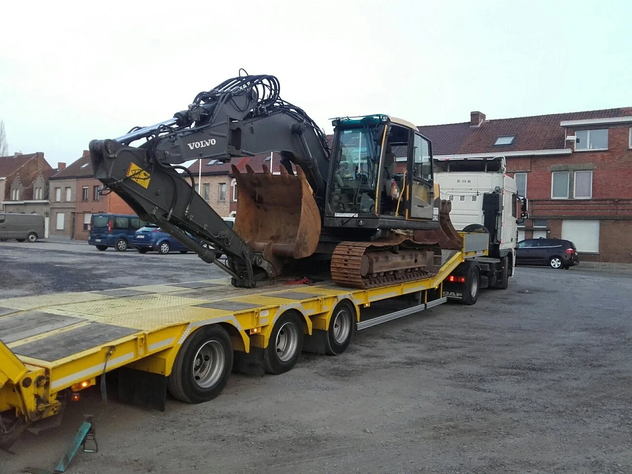 Een grote graafmachine op een gele transportwagen, klaar voor vervoer op een parkeerplaats met huizen op de achtergrond.