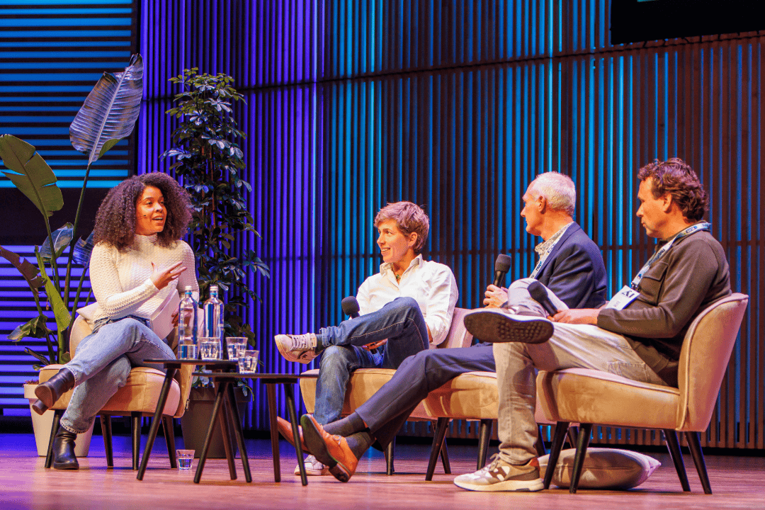 Four people seated on stage having a panel discussion, with a woman speaking and three men listening, in a colorful auditorium setting.