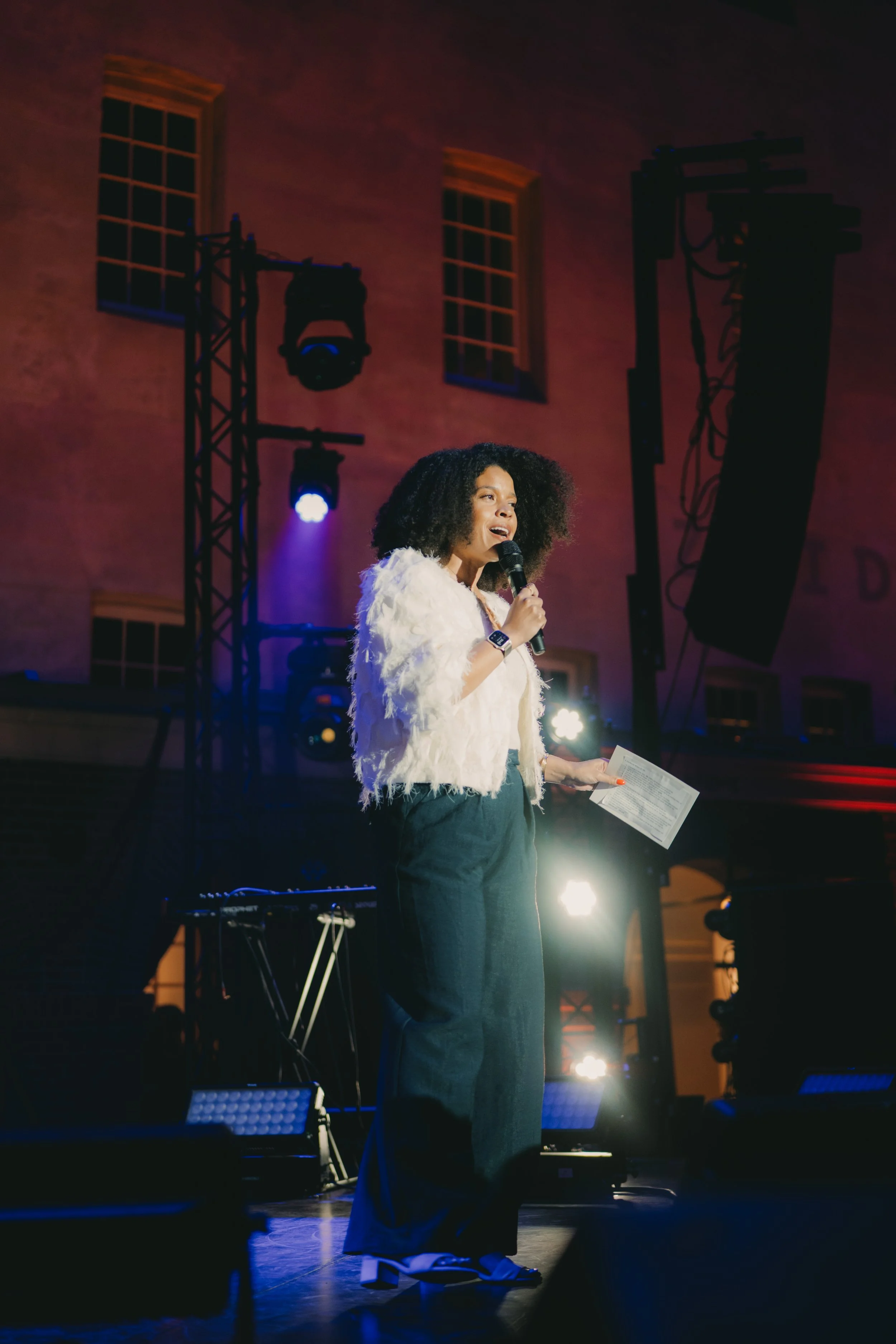 A woman with curly black hair holding a microphone and sheet of paper, performing on stage with stage lighting and sound equipment behind her. It's the Scheepvaartmuseum during SAIL at the event Changing Currents