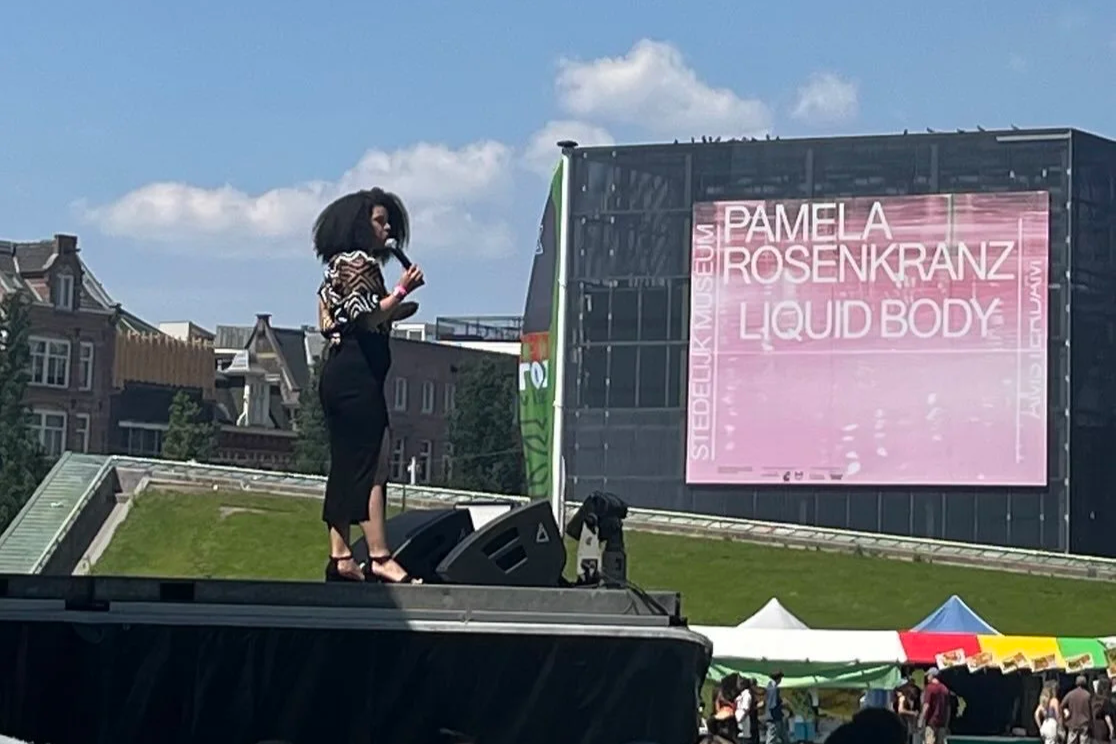A woman standing on an outdoor stage holding a microphone at the Keti Koti festival at Museumplein, Amsterdam in 2025. There are tents and people in the foreground.