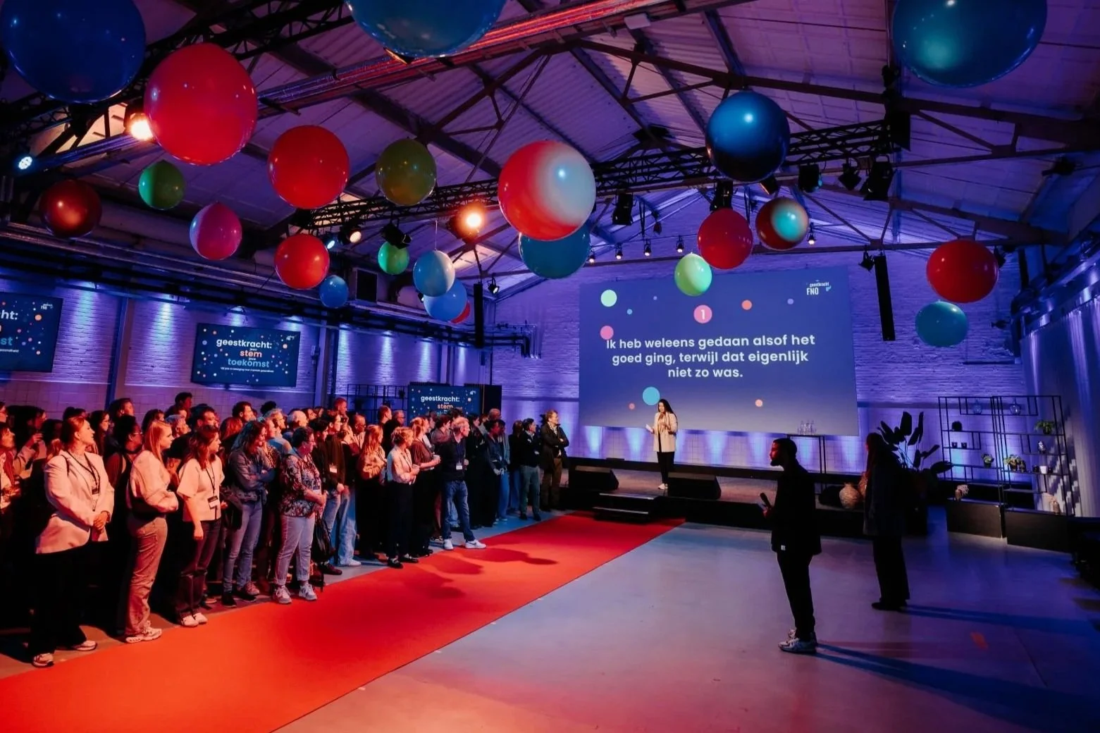 People attending a presentation or conference in a large, decorated venue with colorful balloons hanging from the ceiling and screens displaying text in Dutch. For FNO Geestkracht. 