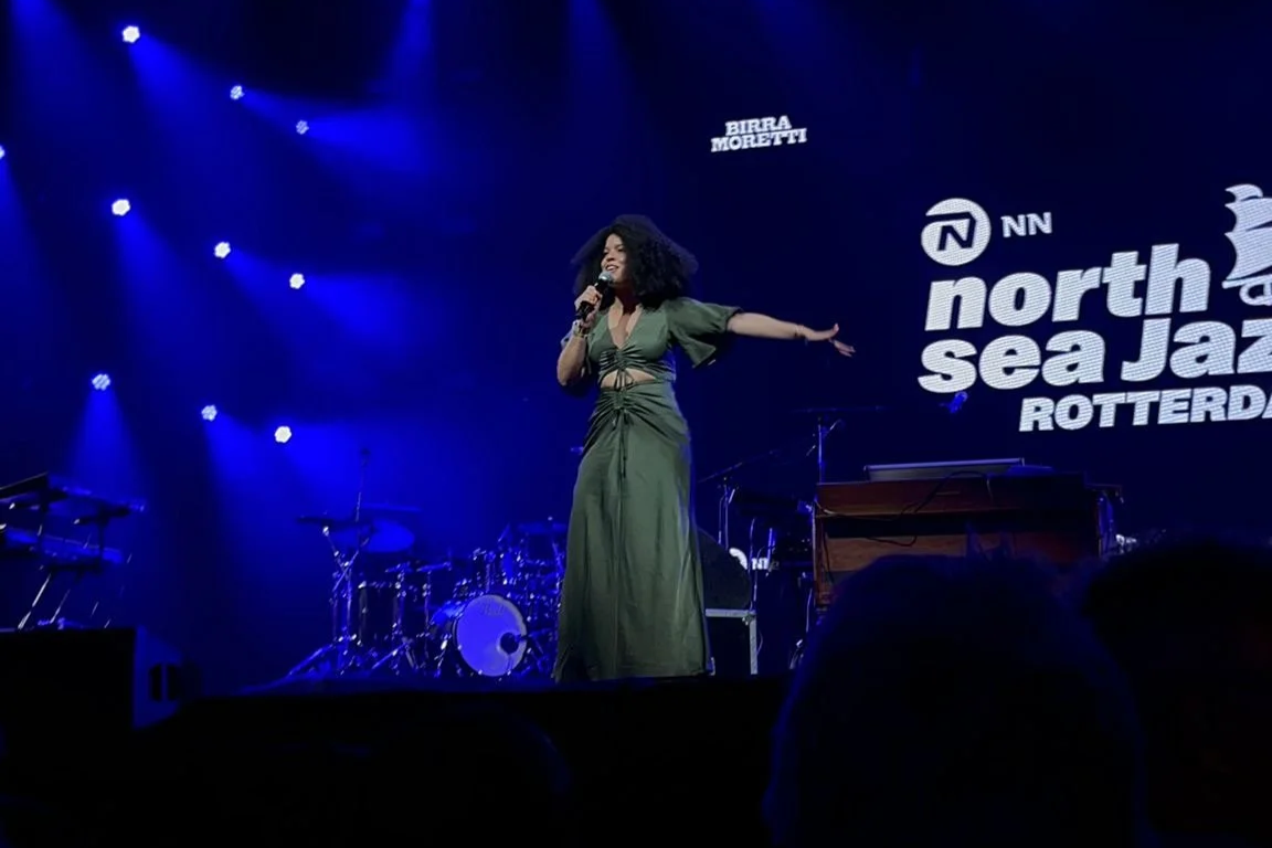 Jazz singer performing on stage at North Sea Jazz Festival Rotterdam, with blue stage lights, drums, and a keyboard in the background.