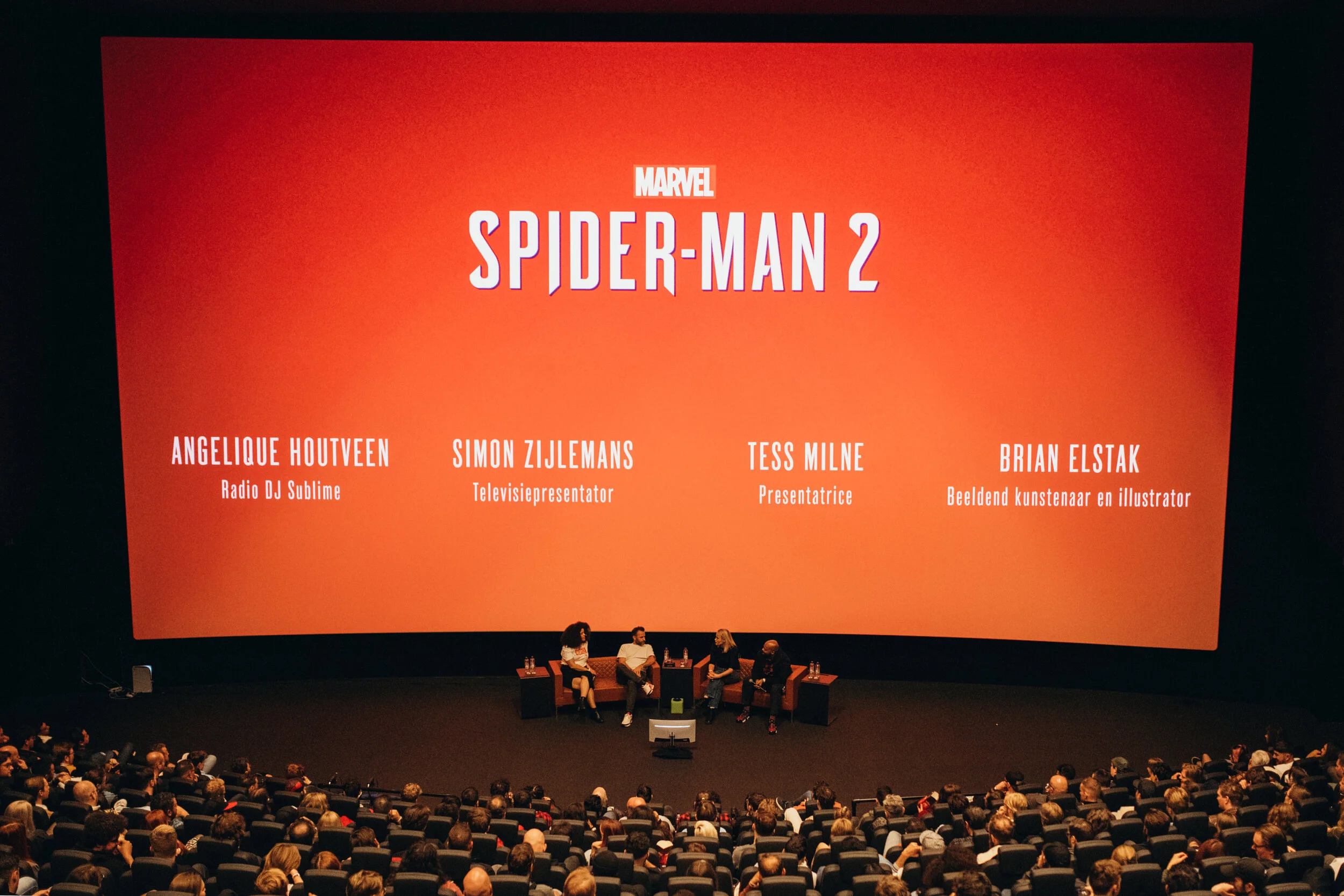 Presentation event for Marvel's Spider-Man 2 with four panelists on stage, sitting in chairs with microphones and water bottles, in front of a large orange-red screen displaying the event's title and panelists' names and roles, surrounded by an audience.