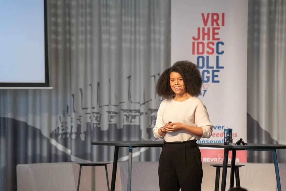 Young woman giving a presentation at a conference with a screen and banner in the background.