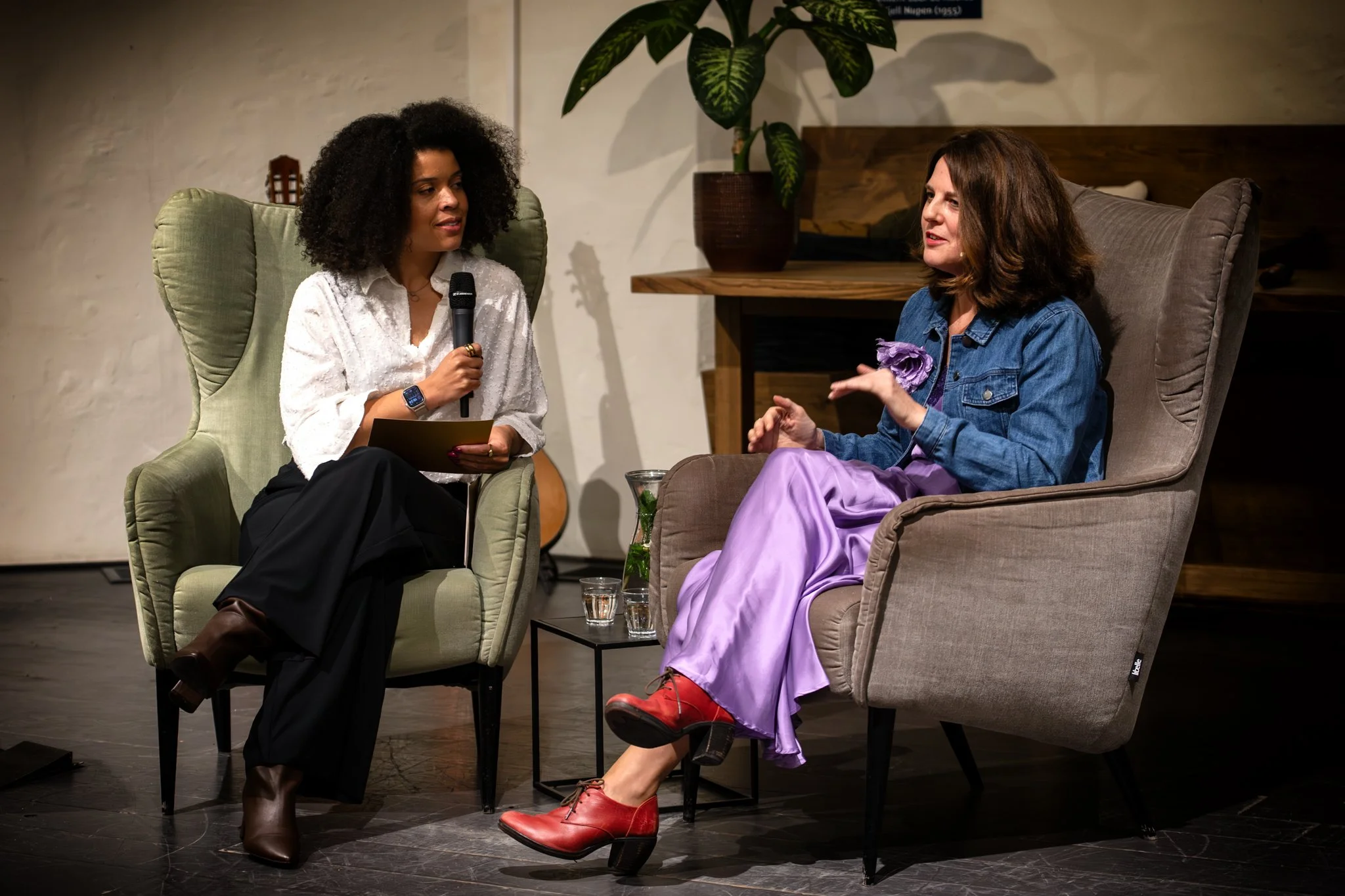 Two women having a conversation in a comfortable setting. One woman, with curly black hair, is sitting in a green armchair, holding a microphone and a notebook. The other woman, with shoulder-length brown hair, is sitting in a beige armchair, wearing a denim jacket and purple skirt, gesturing with her hand. 