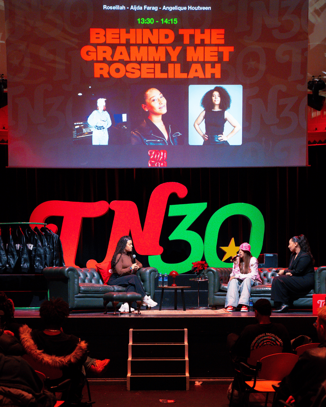 Stage with three women participating in a discussion at Topcon. Large screen displays a presentation titled "Behind the Grammy met Rosellah" with images of Rosellah, and text with names and a schedule. Audience seated watching.