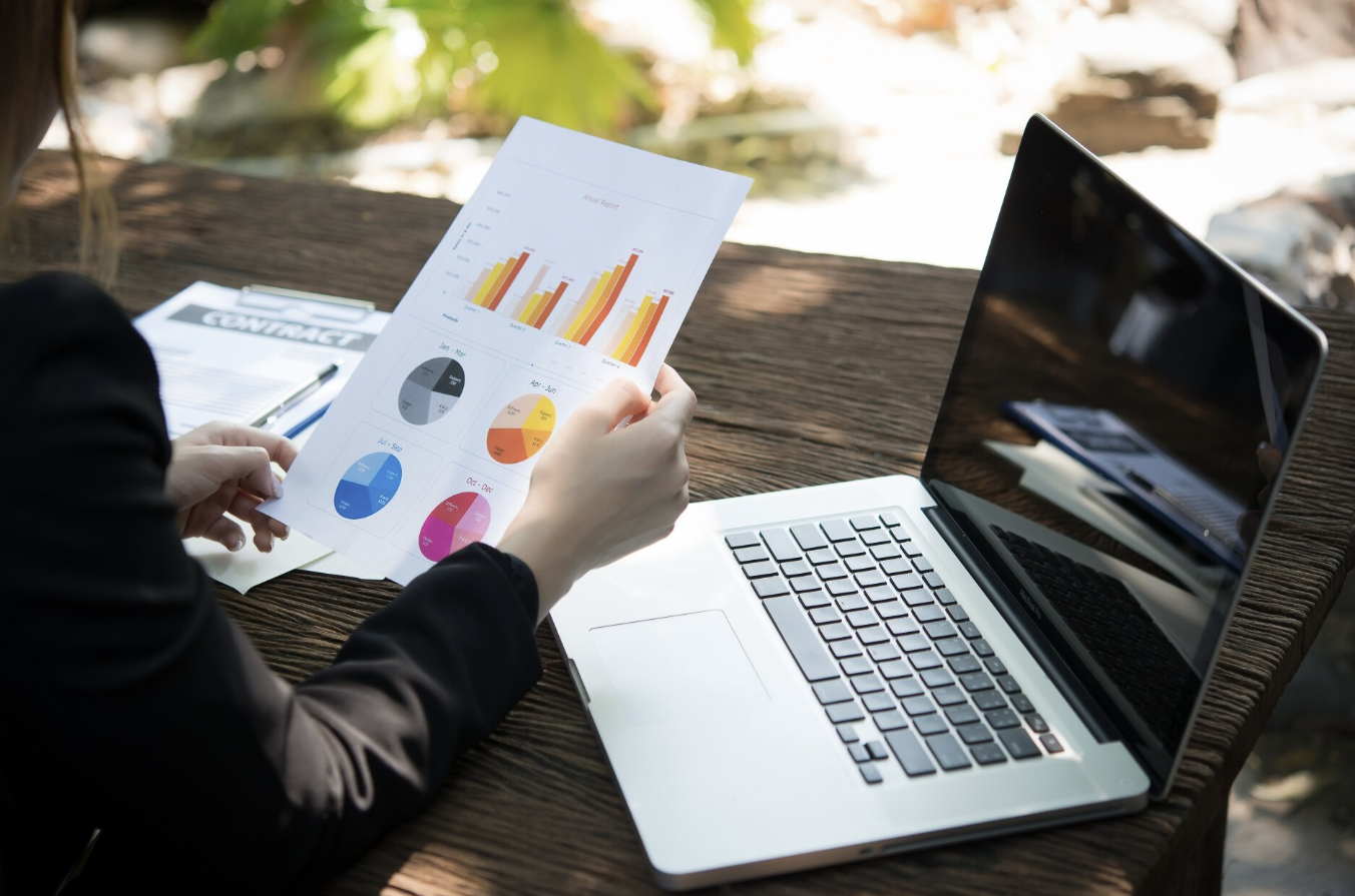 Person holding a printed report with bar and pie charts, sitting at a wooden table with a laptop and a contact book outdoors.