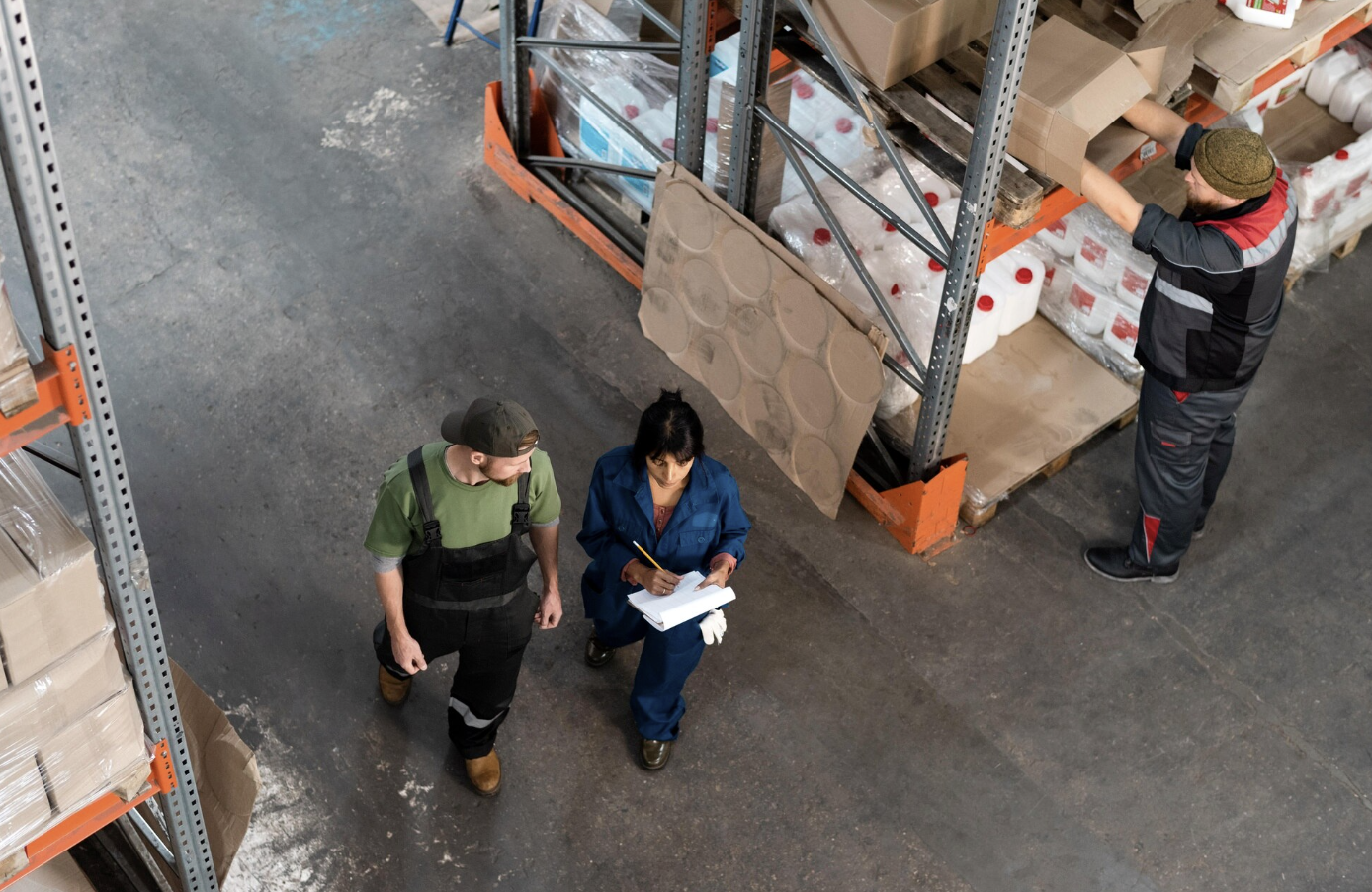 Warehouse workers inspecting inventory, one taking notes while another reaches for items on high shelves.