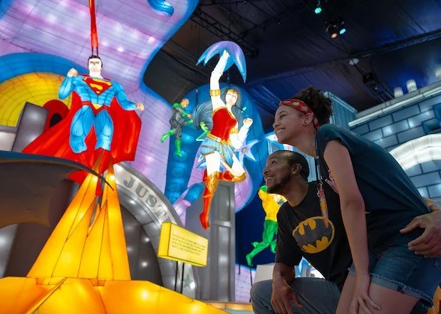 Two people, a man wearing a Batman T-shirt and a woman with curly hair, are smiling and looking at superhero and cartoon character statues illuminated in an indoor amusement park or themed attraction.