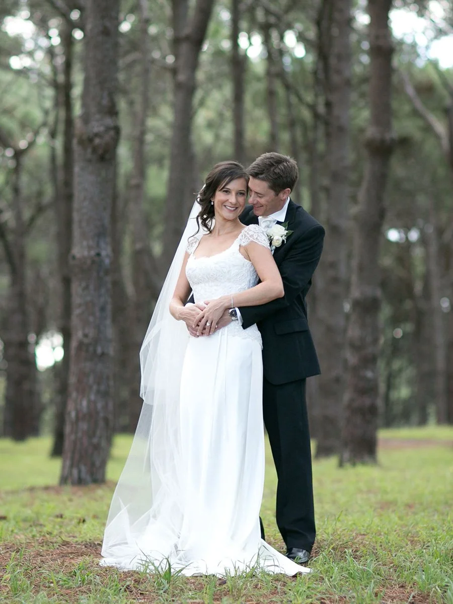 Couple embrace among the pine trees in Centennial Park, foreheads nearly touching.