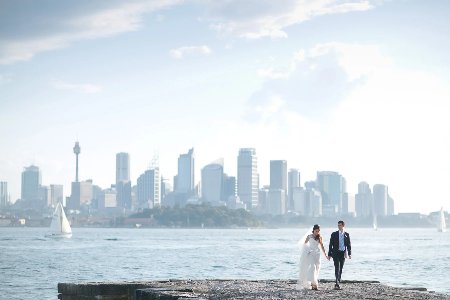 Bride and groom hand in hand on the stone pier with the Sydney city skyline behind, Bradleys Head Mosman