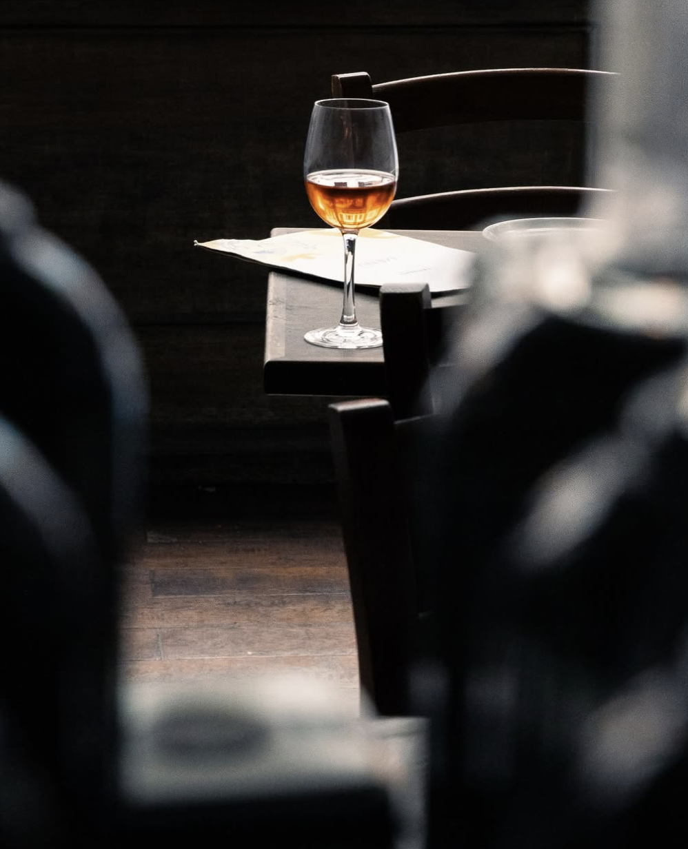 A glass of pink wine on a dark wooden table, with blurred chairs and a dark background.