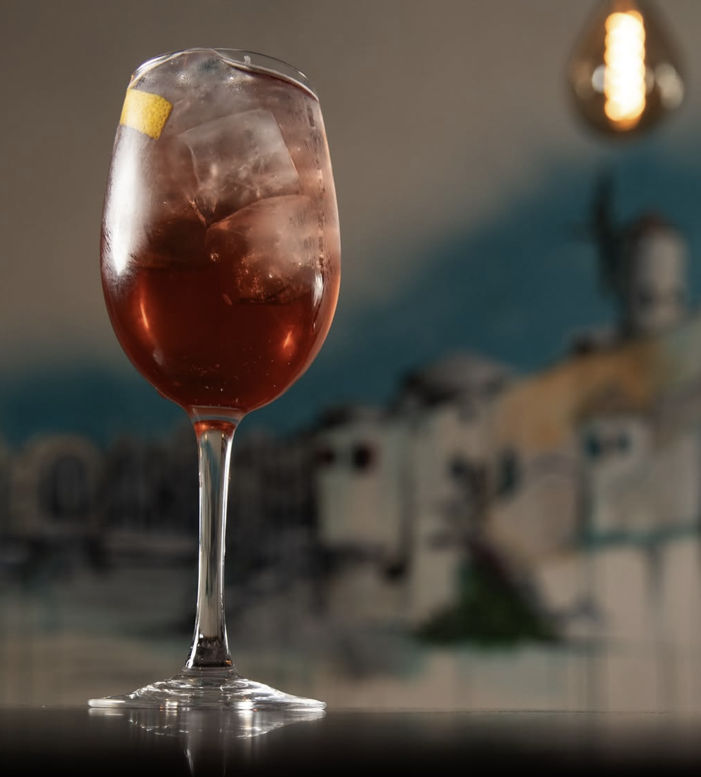A glass of rosé wine with ice cubes and a lemon twist inside, on a table with a blurred background showing a wall and a lamp.