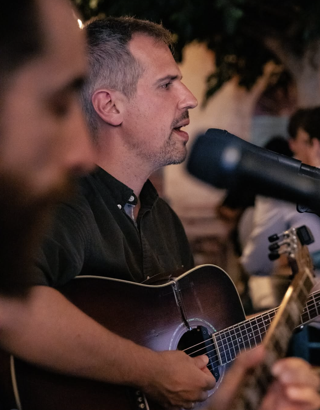 A man singing into a microphone while playing an acoustic guitar at an outdoor event, with blurred background of other people and trees.