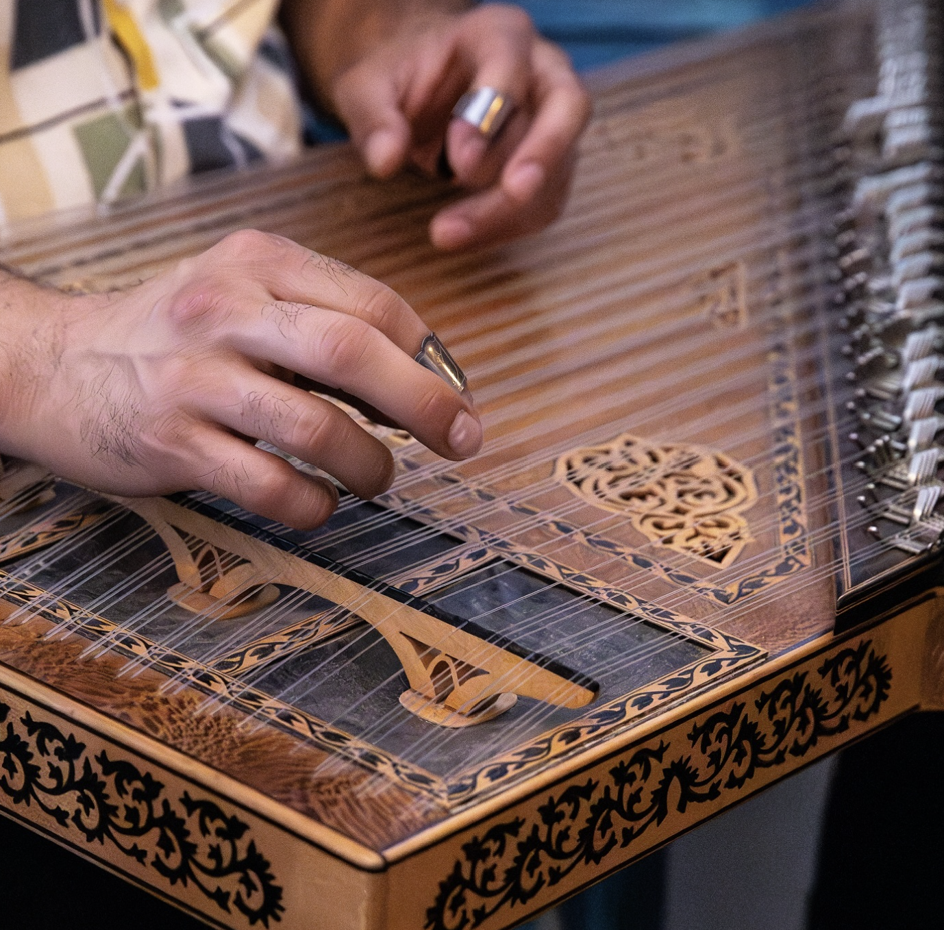 Close-up of hands playing a wooden hammered dulcimer with intricate carvings and metal strings.