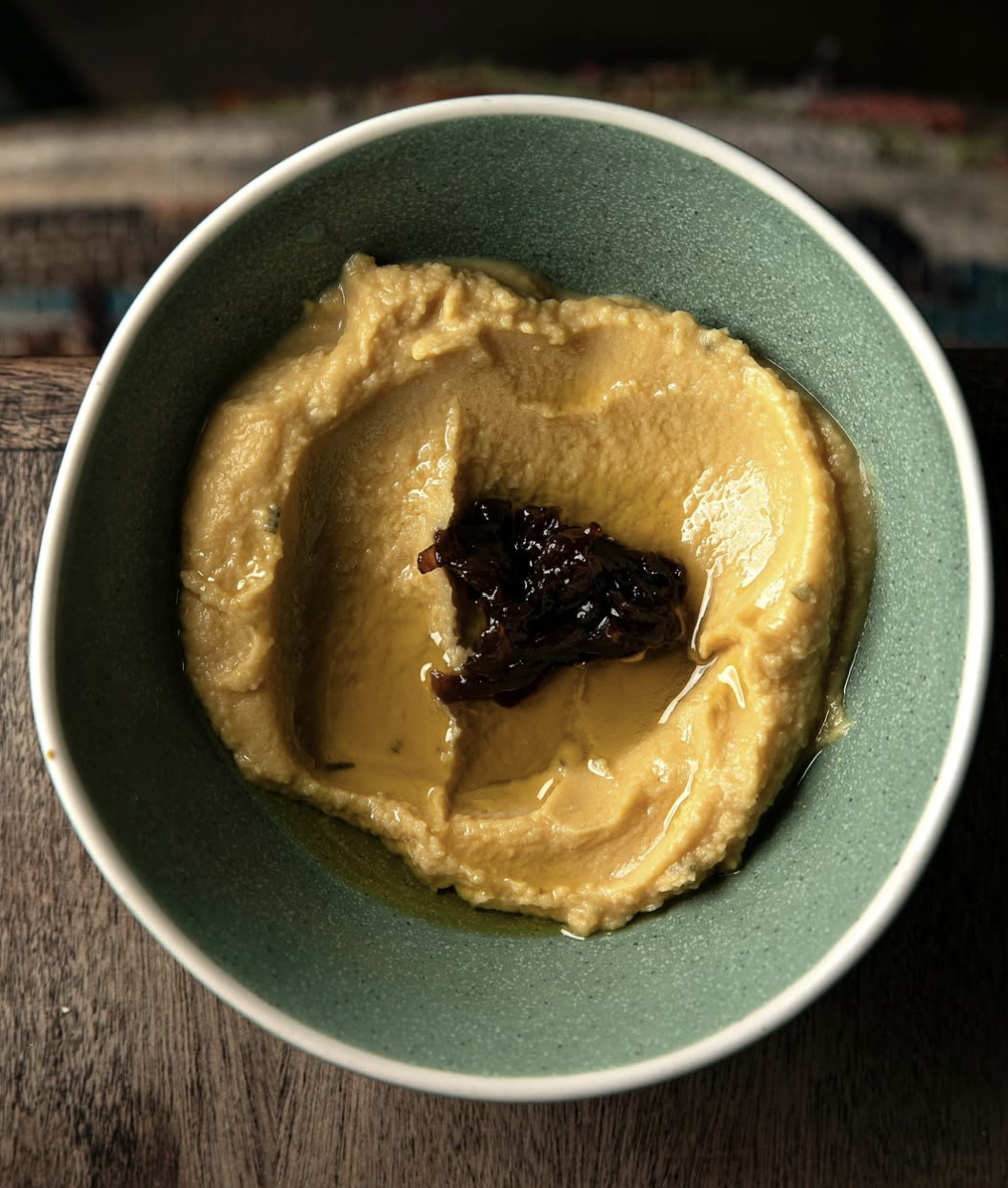 A bowl of creamy hummus topped with dark olive tapenade, placed on a wooden surface.