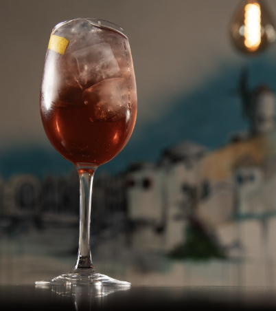 A tall glass of iced dark red beverage with a lemon slice on the rim, set against a blurred background of a coastal town.