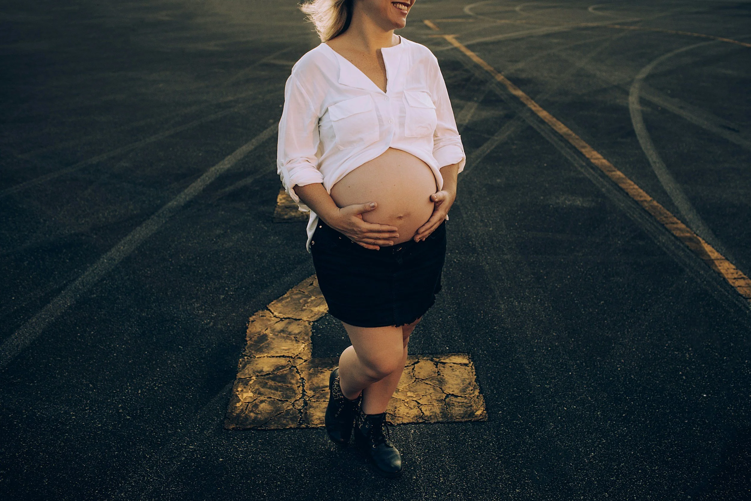 A pregnant woman standing in an empty parking lot, smiling with her hands on her belly, wearing a white shirt and a black skirt.