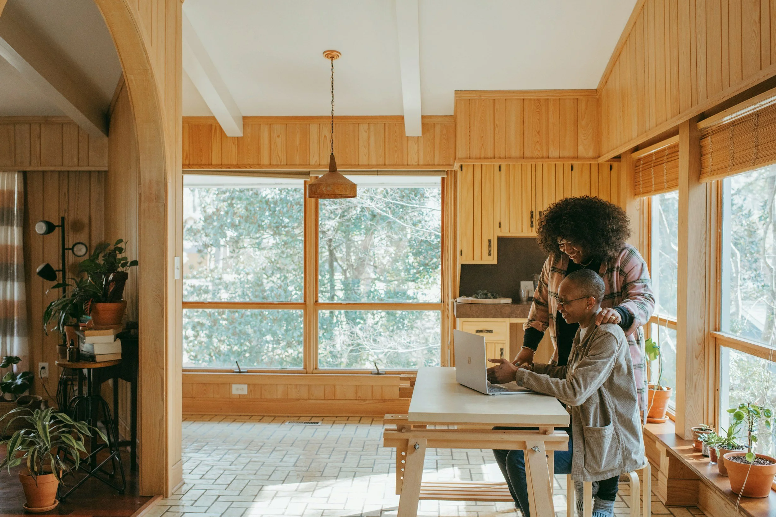 A woman and a young person working on a laptop in a sunlit kitchen with wooden walls and large windows showing trees outside.
