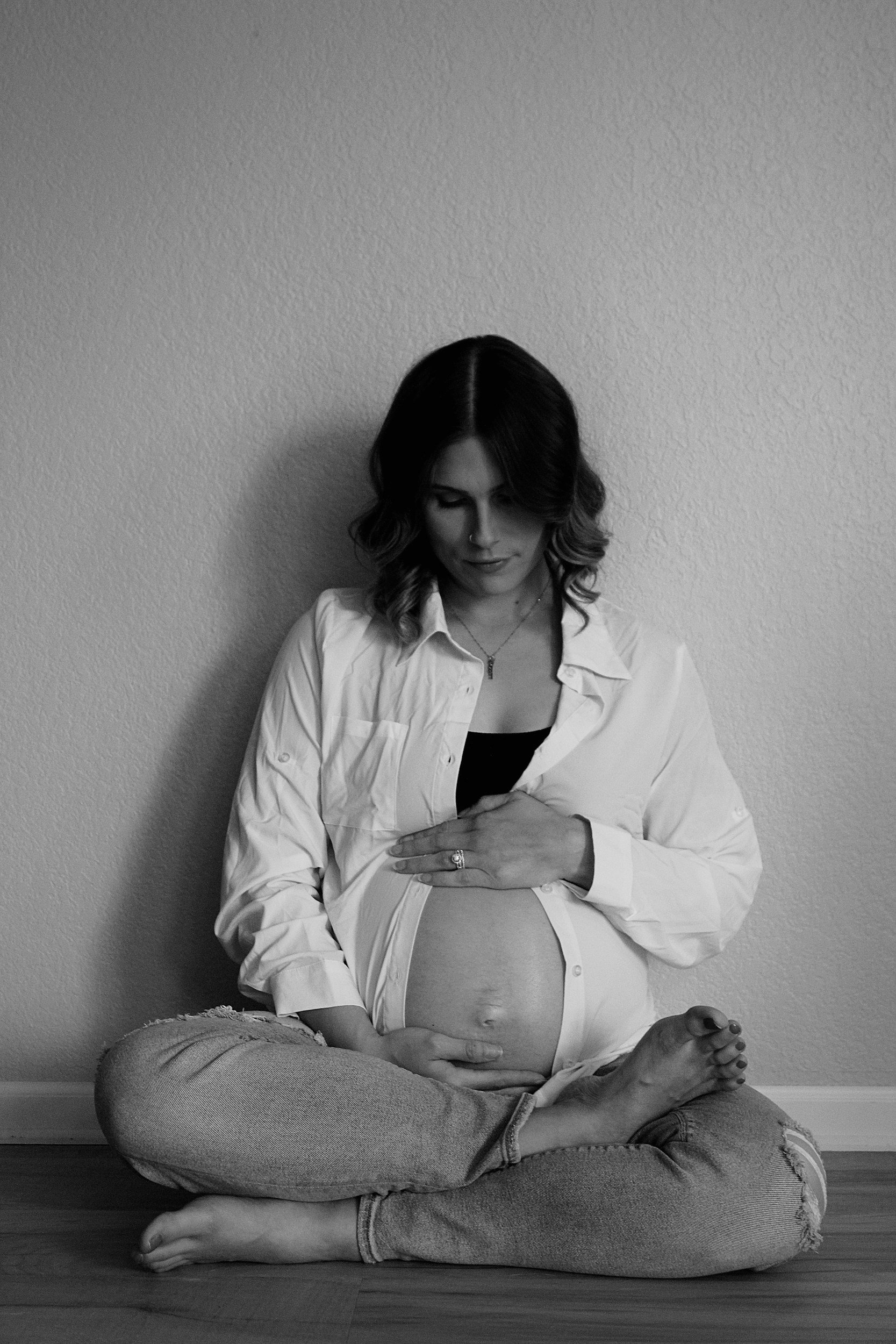 A pregnant woman with dark hair, sitting cross-legged on the floor against a plain wall, wearing a white shirt and ripped jeans, looking down at her belly.