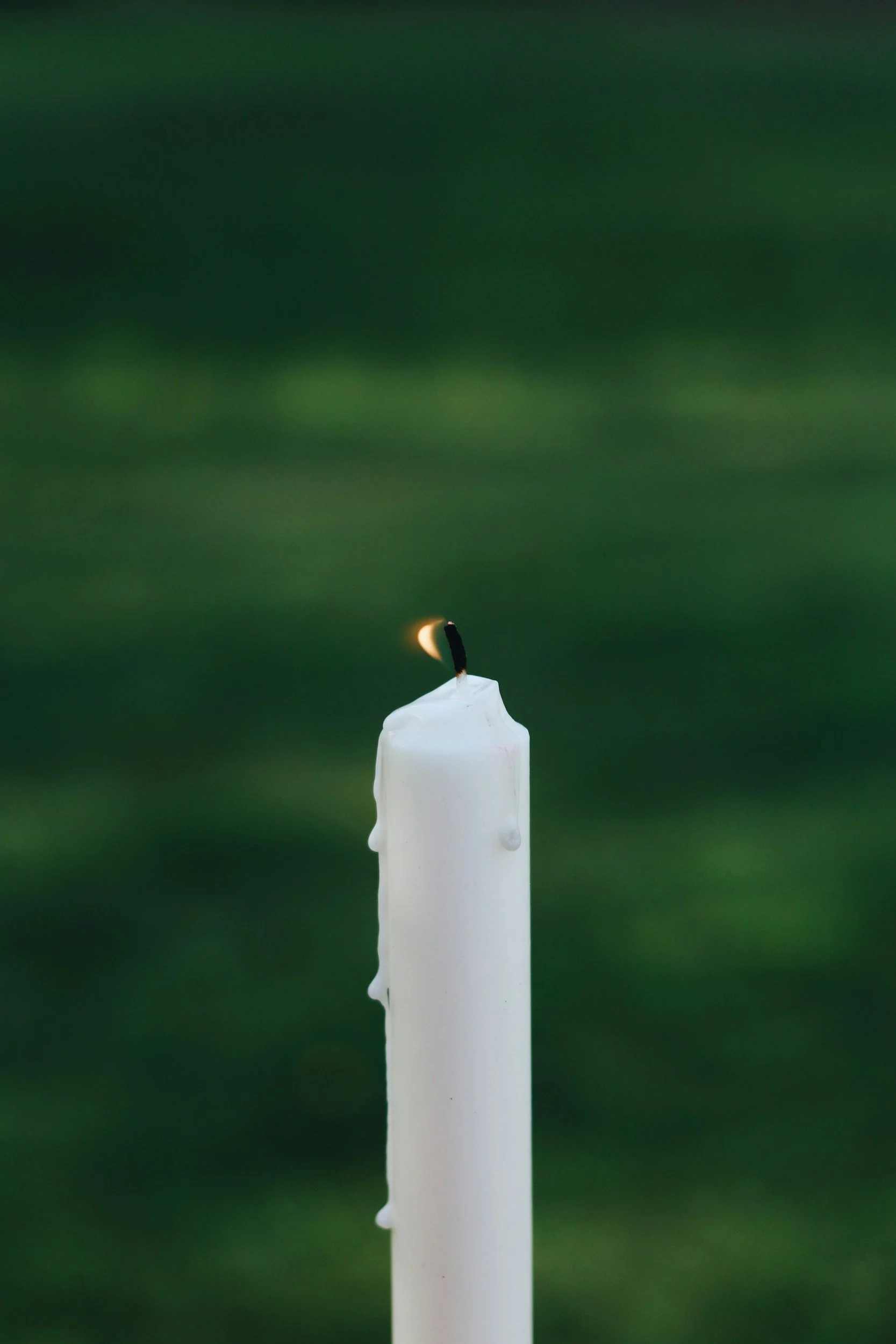 A close-up of a white candle with a burning wick against a dark green blurred background.