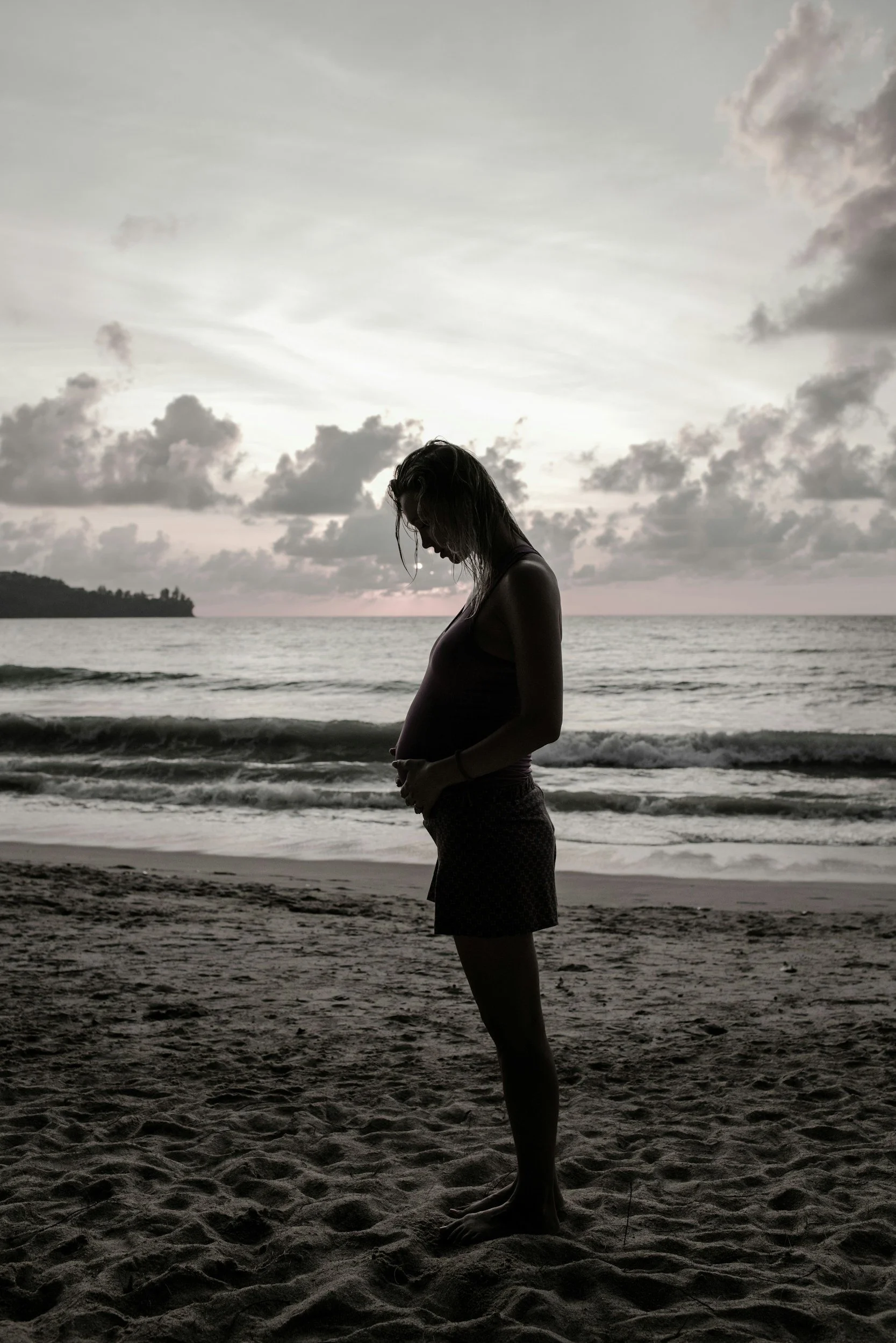 Silhouette of a pregnant woman standing on a beach during sunset with clouds in the sky.