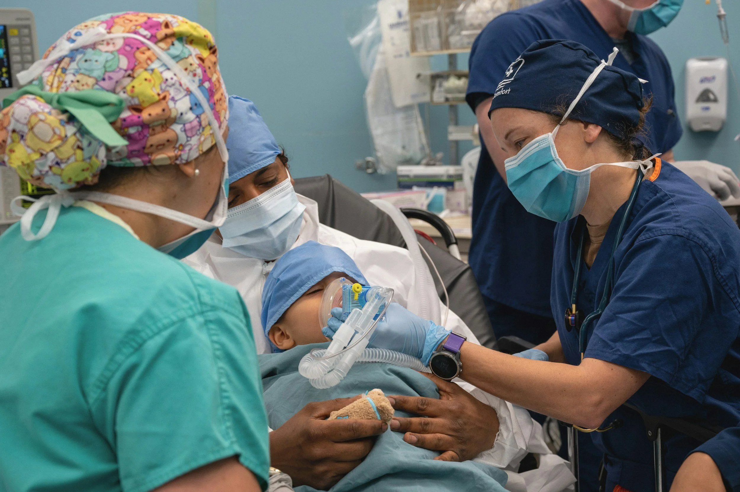 Medical team attending to a young patient in a hospital room. The child is lying in bed with a breathing mask, surrounded by healthcare professionals wearing scrubs, masks, and gloves.