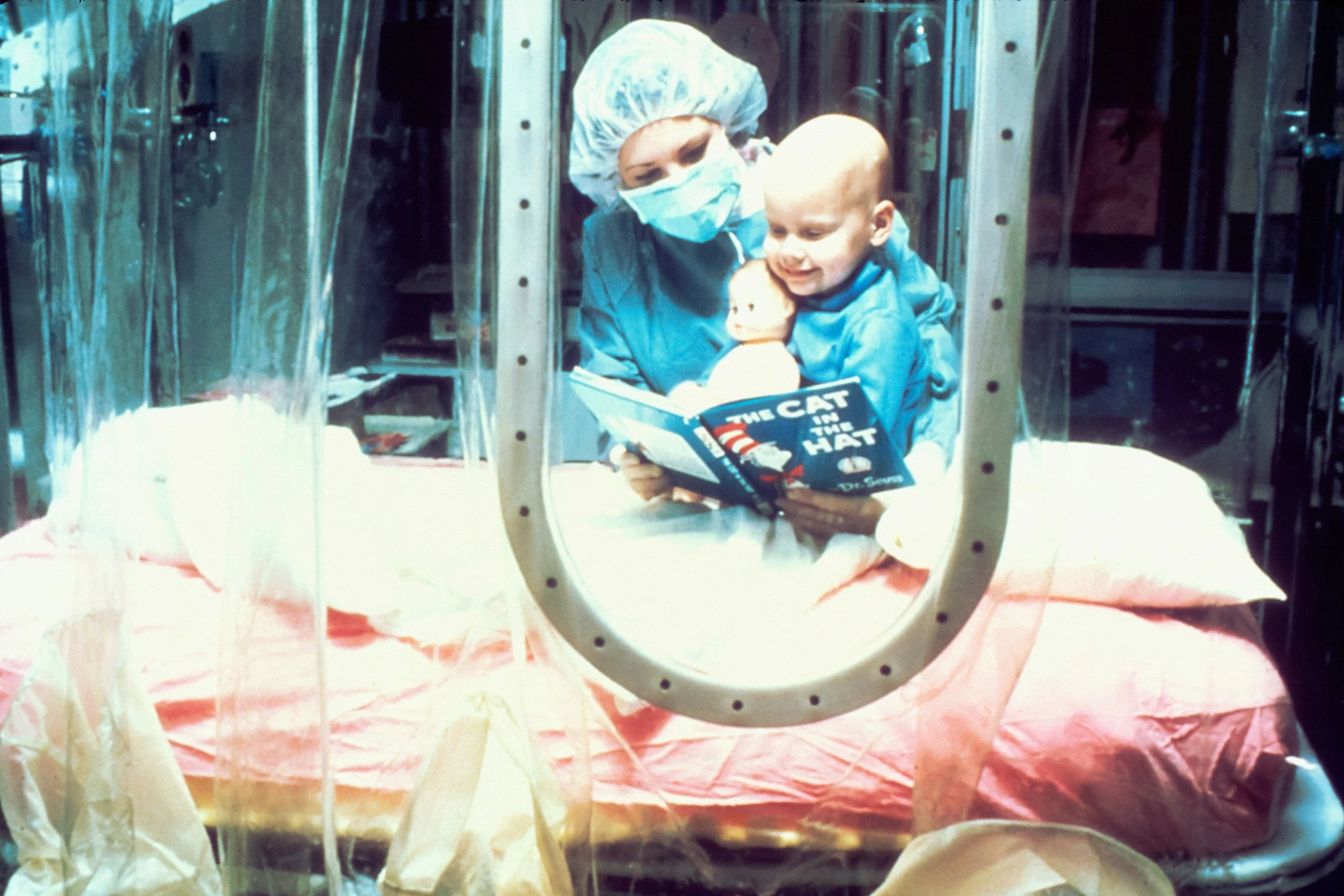 A young child lying in a hospital incubator, holding a doll and reading a book titled 'The Cat in the Hat.' She is wearing a surgical cap and mask, with a healthcare worker beside her also wearing scrubs.