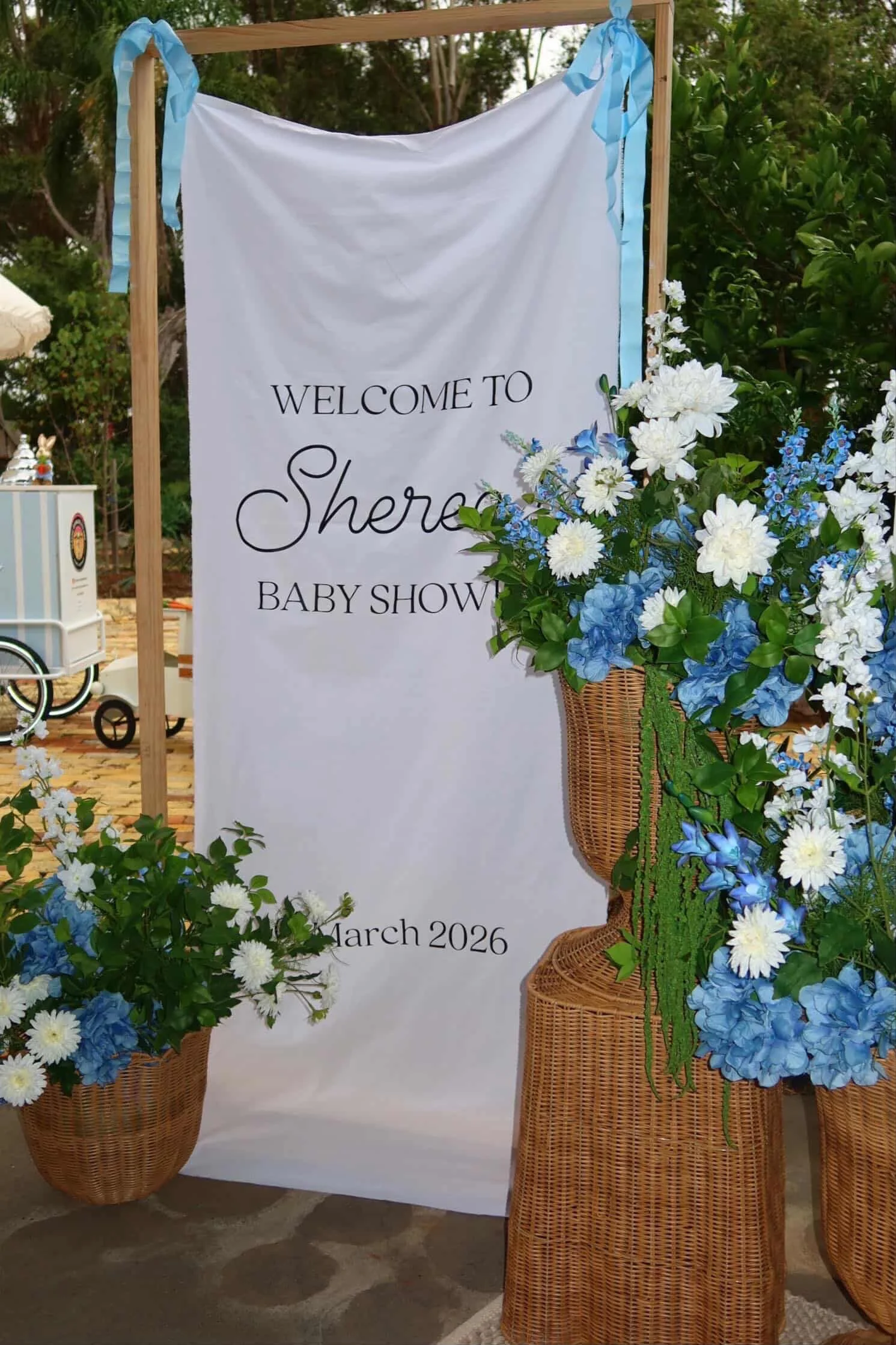 Decorative welcome sign for a baby shower with white and blue flowers in wicker baskets, and blue ribbons on a wooden frame, set outdoors.