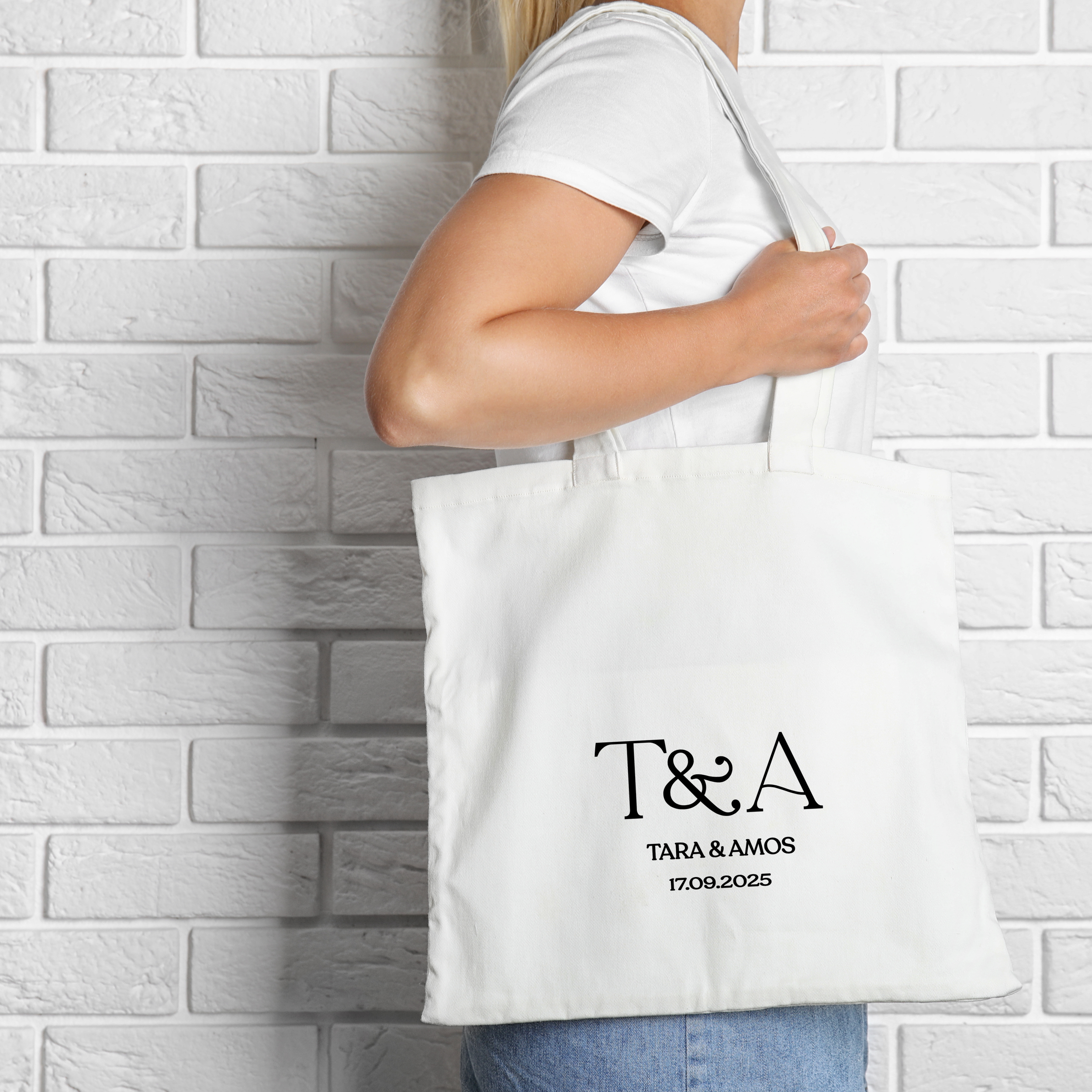 A woman holding a white tote bag with black text that reads 'T&A' and additional details 'Tara & Amos' with a date '17.09.2025', standing against a white brick wall.