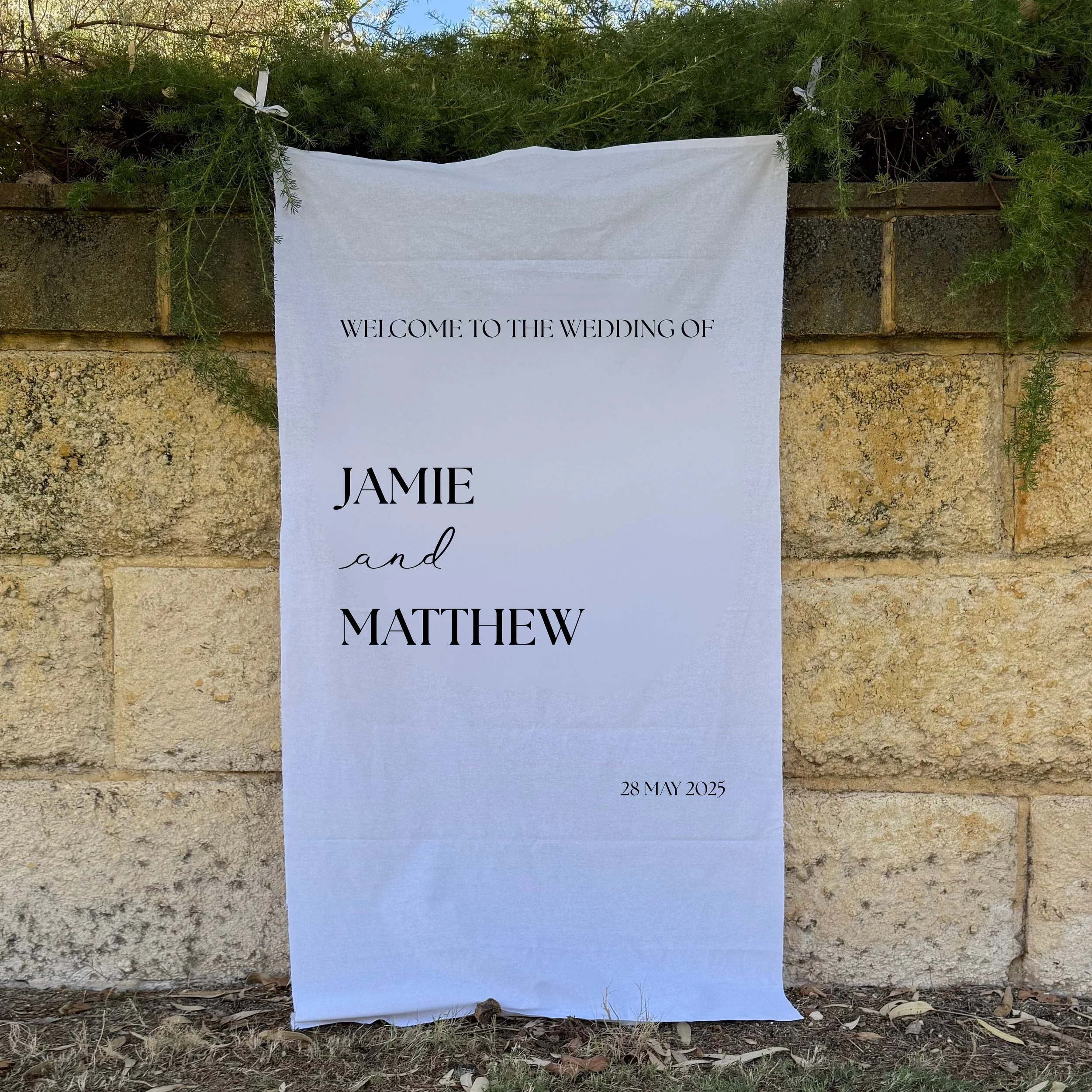 Wedding welcome sign with text 'Welcome to the wedding of Jamie and Matthew, 28 May 2025' hanging on a stone wall, decorated with greenery.