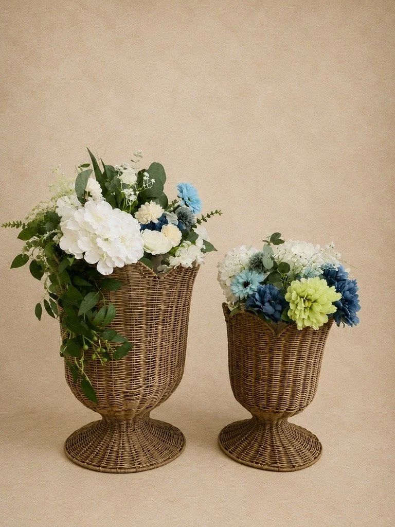 Two woven wicker vases containing white, blue, green, and cream artificial flowers and greenery, against a plain beige background.