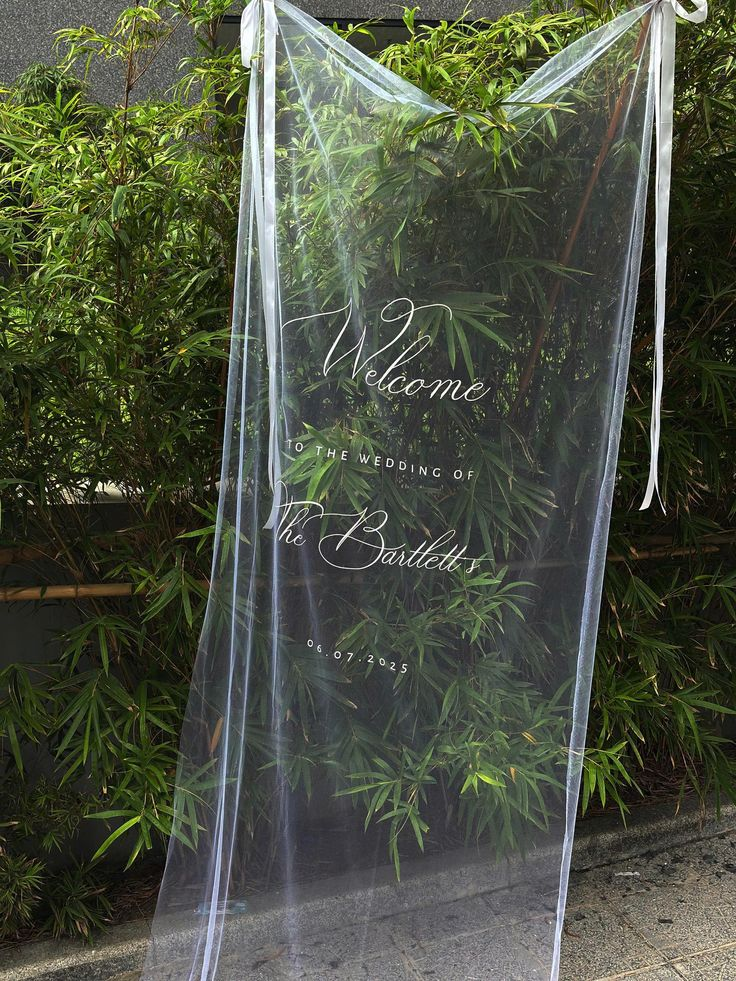 Transparent wedding welcome sign with elegant white cursive text on a background of green plants.