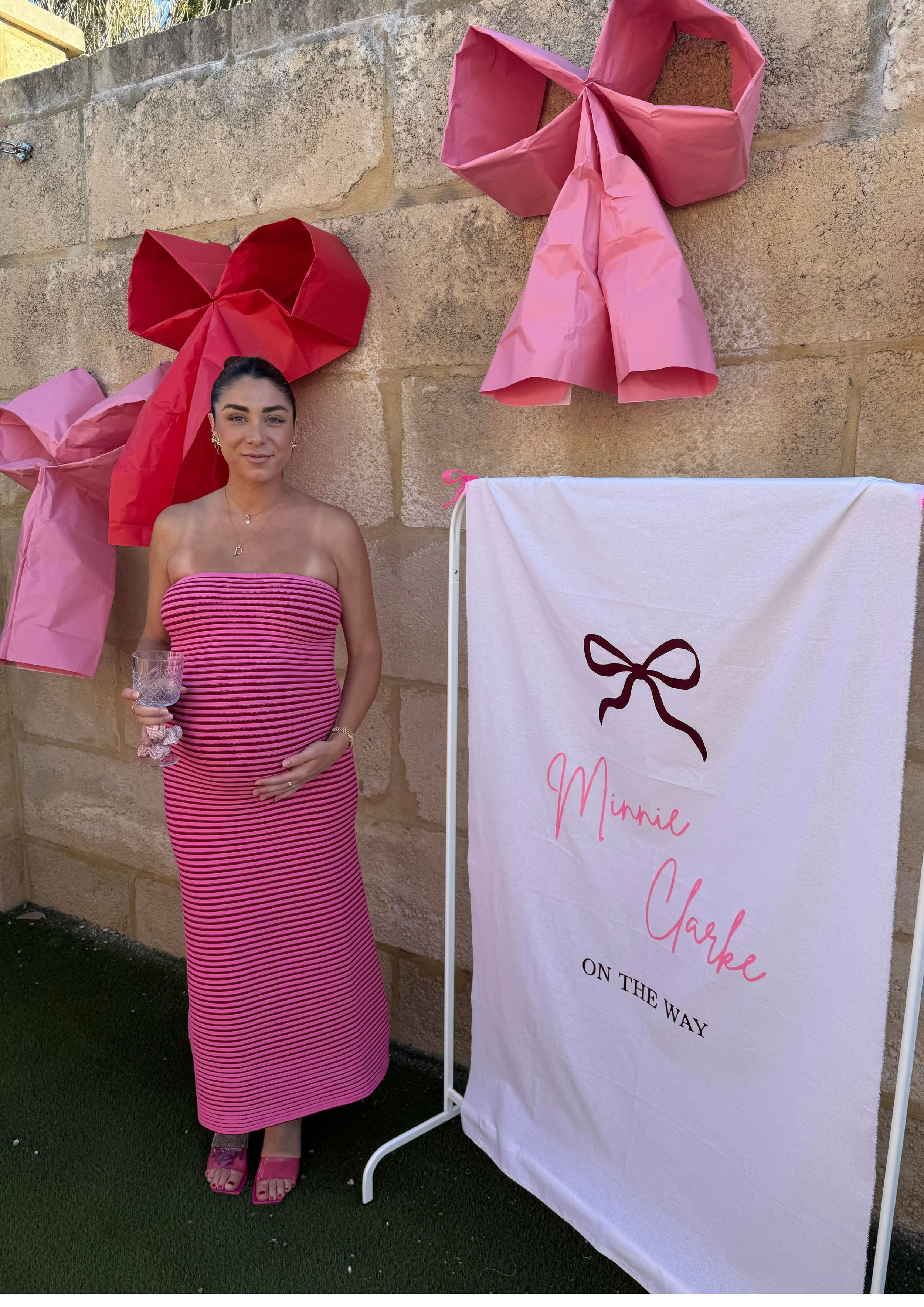 A woman pregnant with dark hair styled in a bun, wearing a pink and black striped strapless dress and pink sandals, standing outdoors in front of a stone wall decorated with large pink and red paper bows. She is holding a glass of water and smiling at the camera. There is a white banner with a black bow graphic and pink cursive text that reads 'Minnie & Carke' and partial text 'ON THE WAY'.