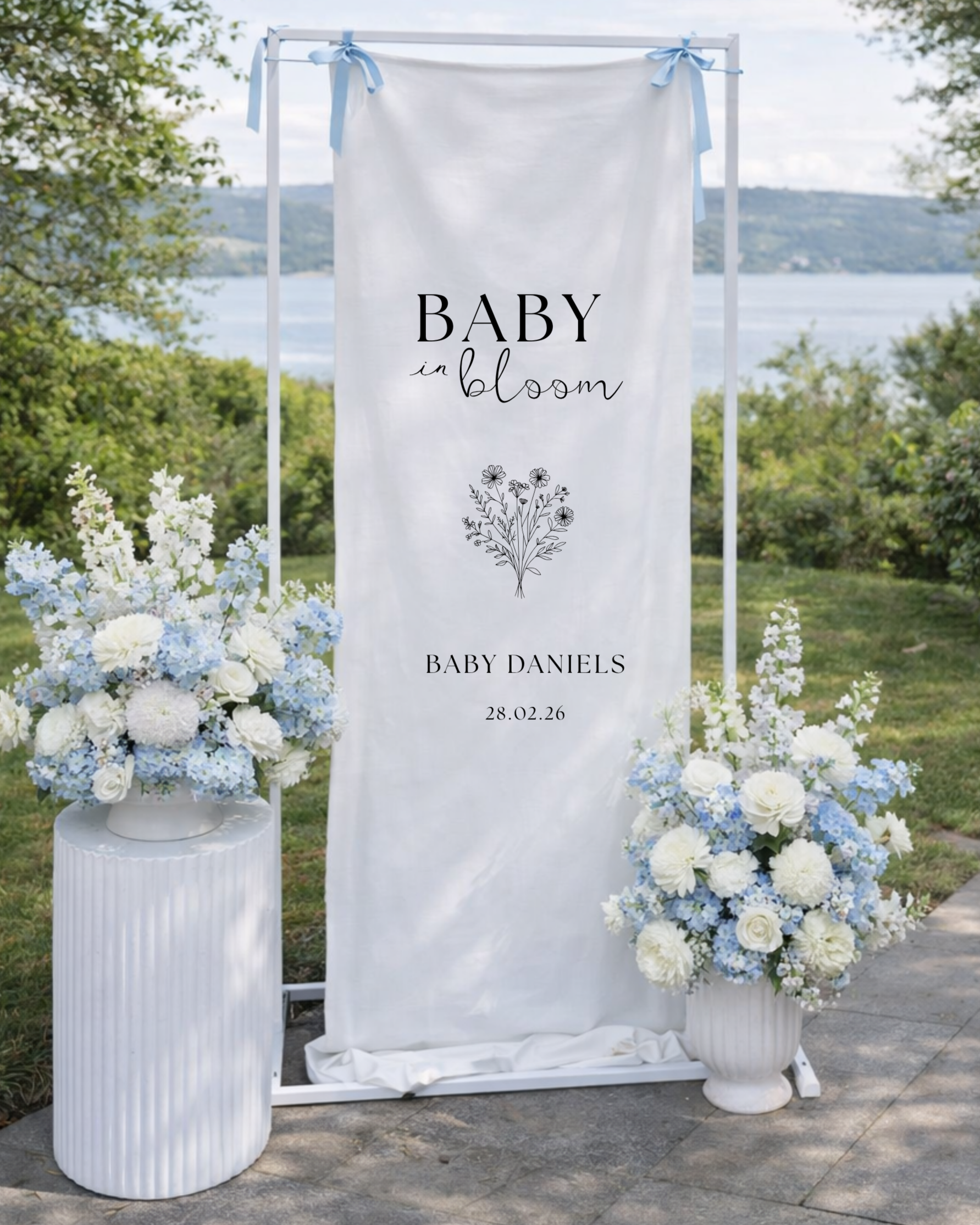 Outdoor wedding display with white flowers and a white banner that reads 'Baby in bloom' with a smaller text 'Baby Daniels' and date '28.02.26', set against a scenic lakeside background.