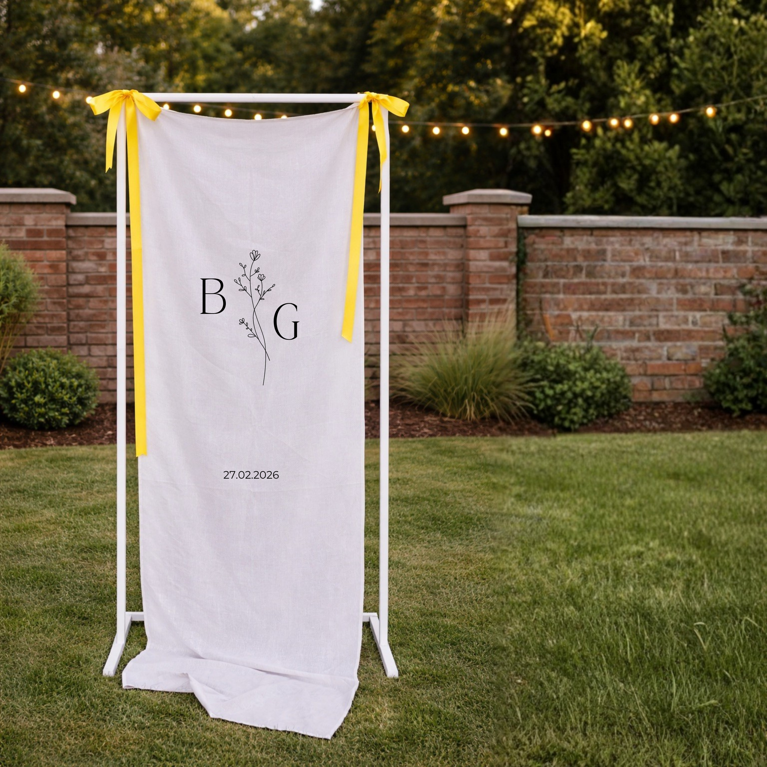 Outdoor wedding or event backdrop with white fabric, yellow ribbons, and string lights, set against a brick wall and greenery