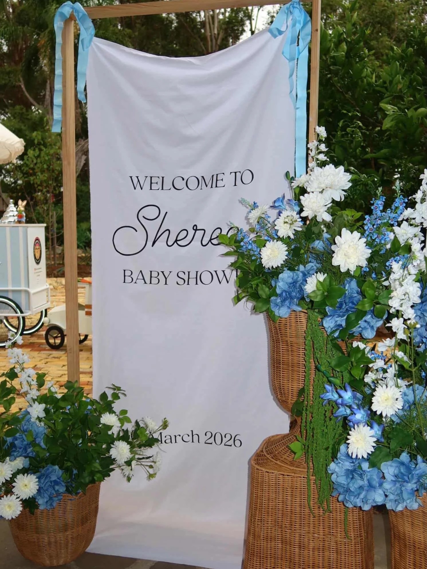 Sign welcoming guests to a Sherer baby shower in March 2026, decorated with blue and white flowers and surrounded by wicker baskets and greenery.