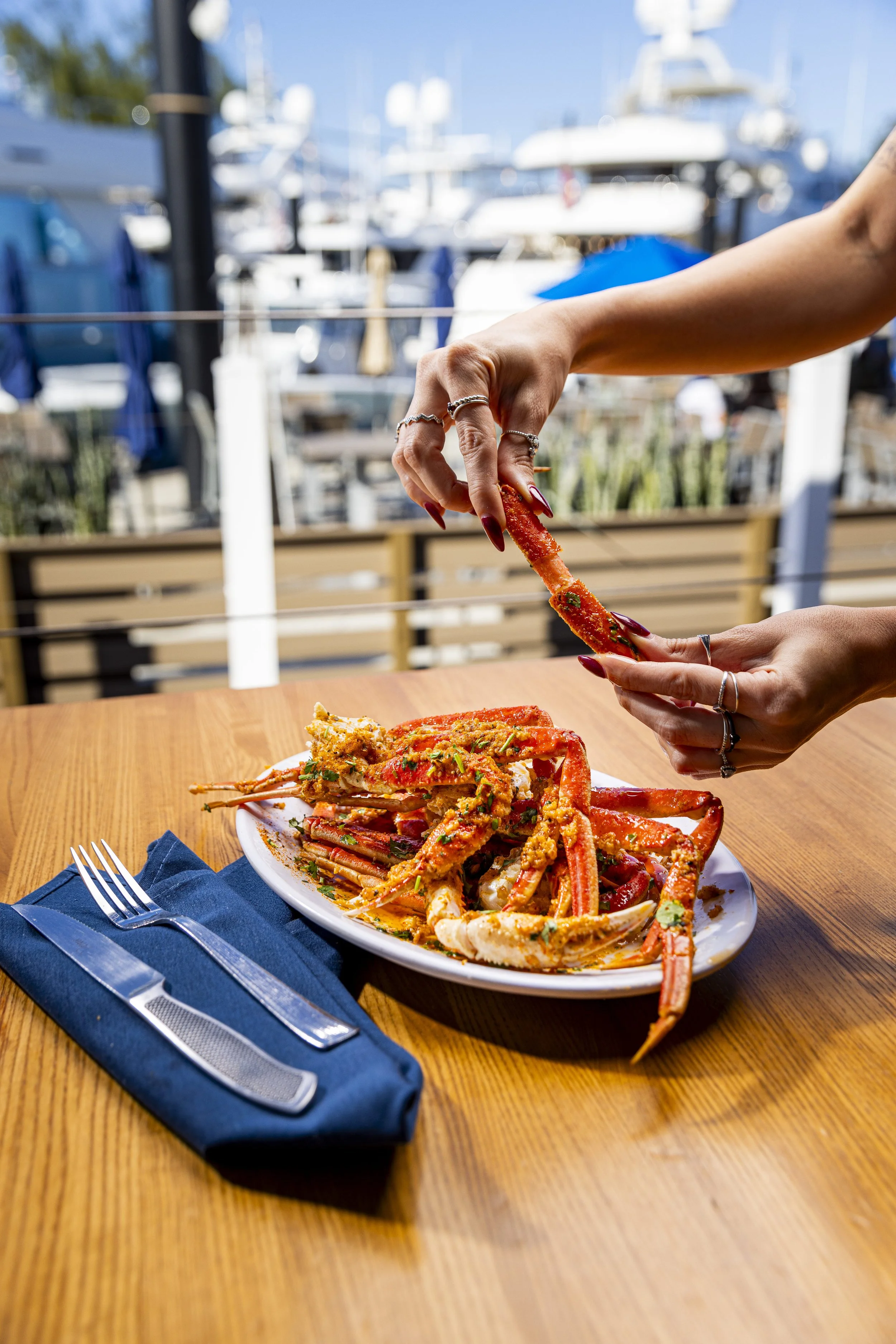 Person's hands with rings picking up a crab leg from a plate of crab legs and seafood, at an outdoor restaurant near a marina.