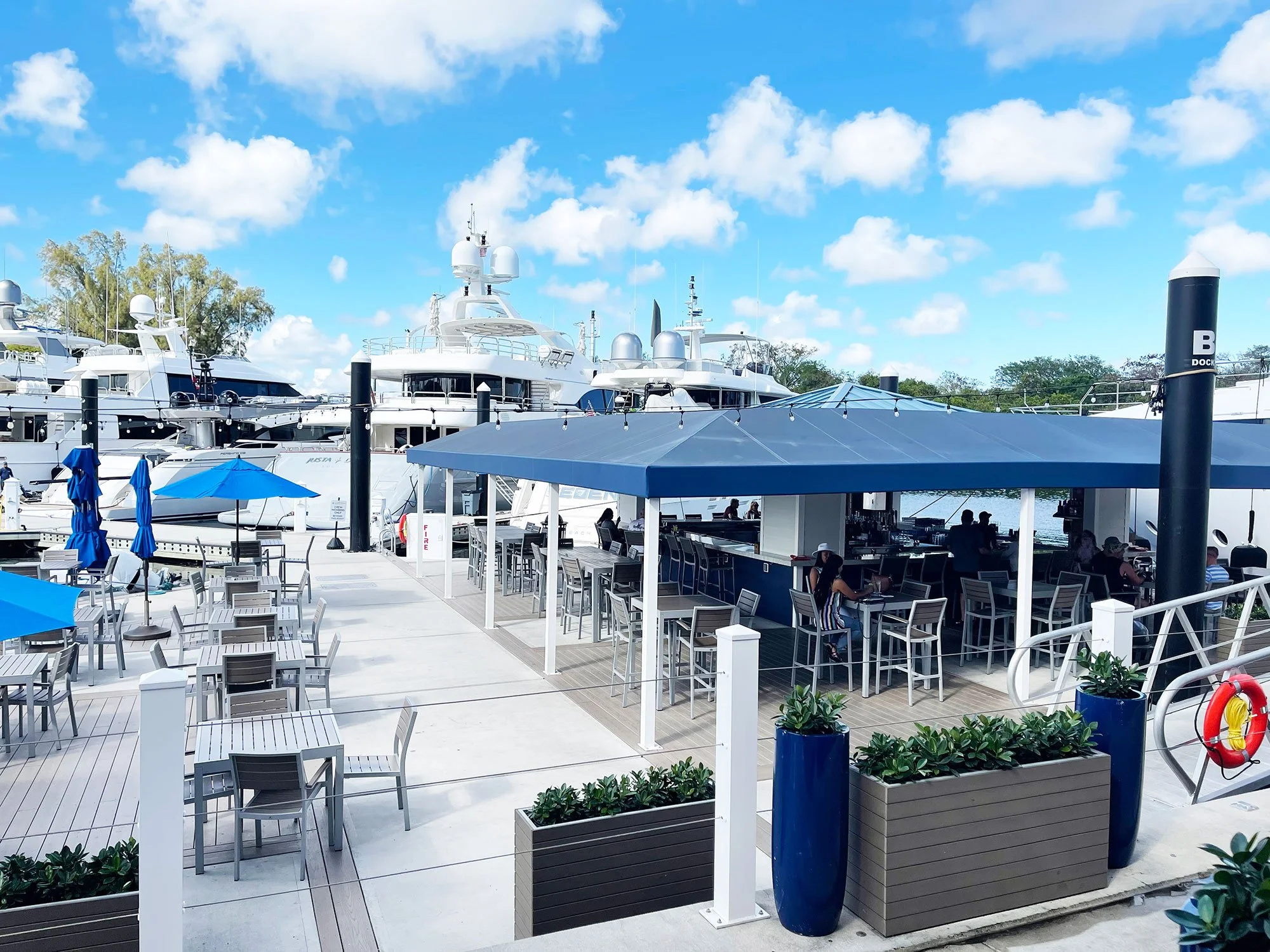 Outdoor dining area at YOT Restaurant overlooking a marina with yachts, featuring patio seating and a covered bar.